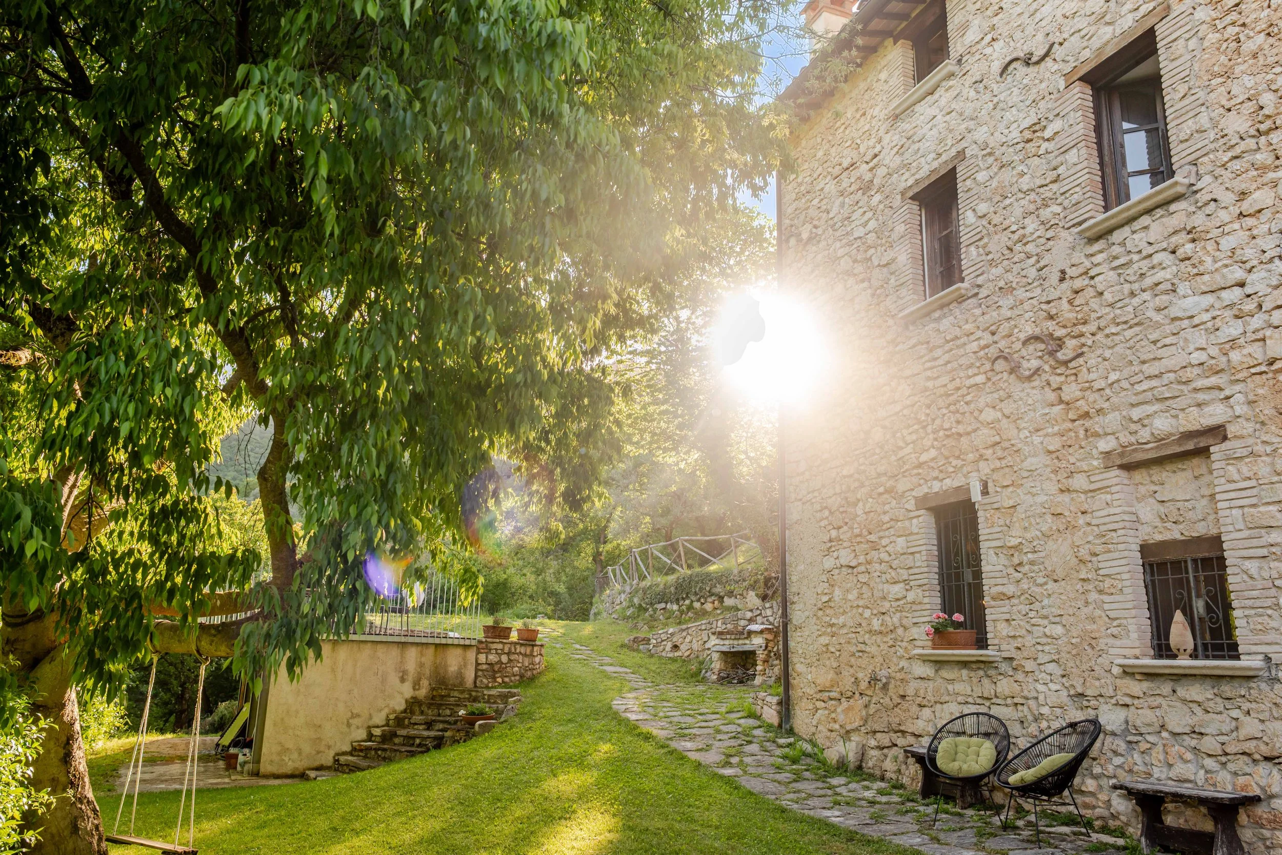 Sunlight shining on a rustic stone house with a lush green garden and outdoor seating.