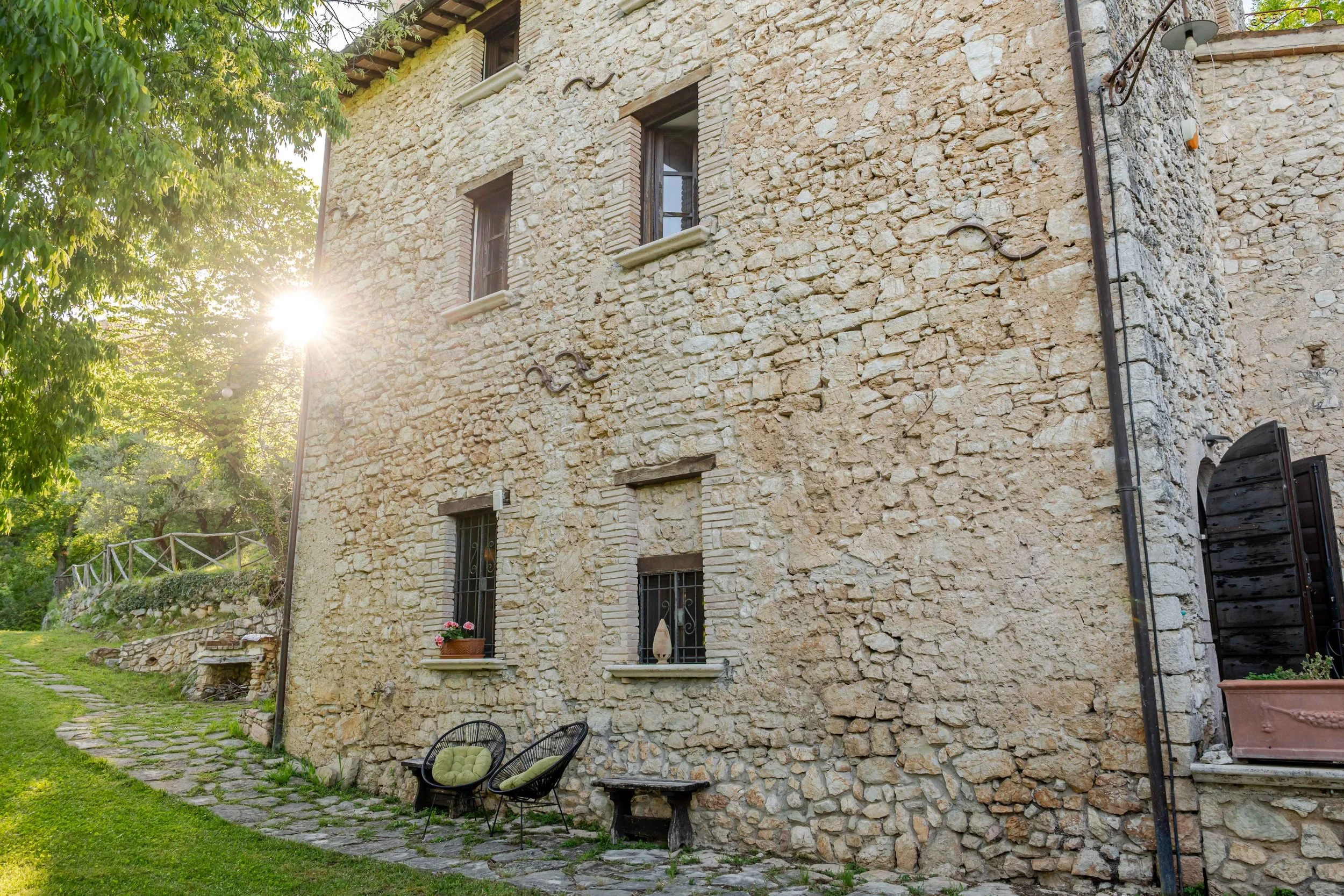 Stone house with small windows, outdoor seating, and surrounded by trees and greenery, sunlight shining from the left.