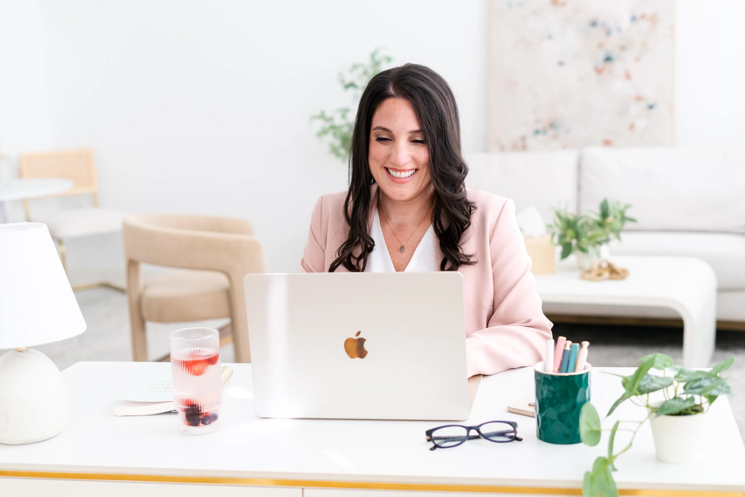 A woman with long dark hair, smiling, wearing a light pink blazer and white shirt, working on a laptop at a white desk with glasses, a glass of red beverage, and various office supplies in a bright, modern room with plants and artwork.