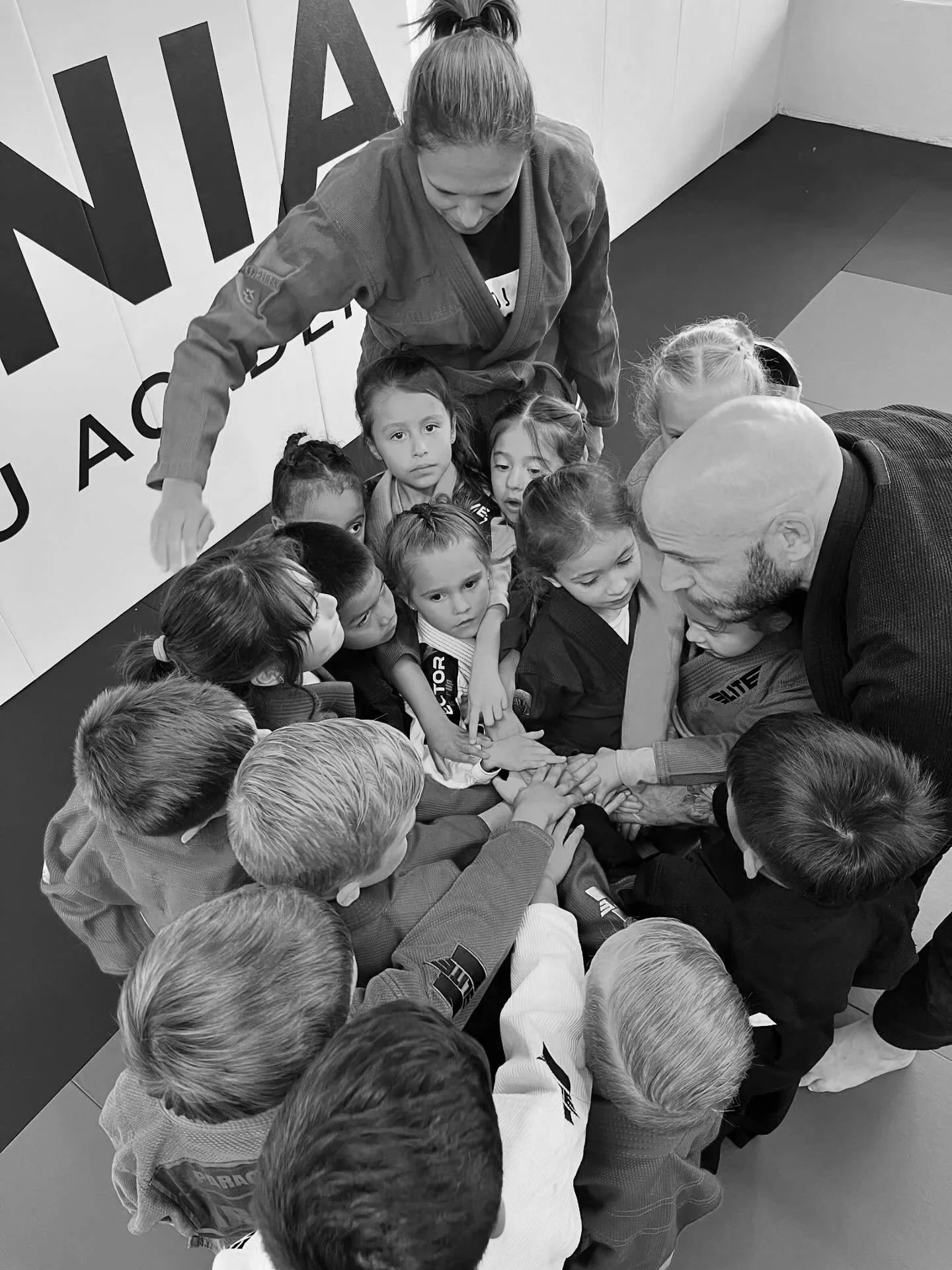 A group of children in Brazilian jiujitsu gis and the instructor in a gi gathered together in a circle, placing their hands in the center for a team cheer or motivational gesture, inside a dojo with mats and a wall with Brazilian jiu-jitsu branding.
