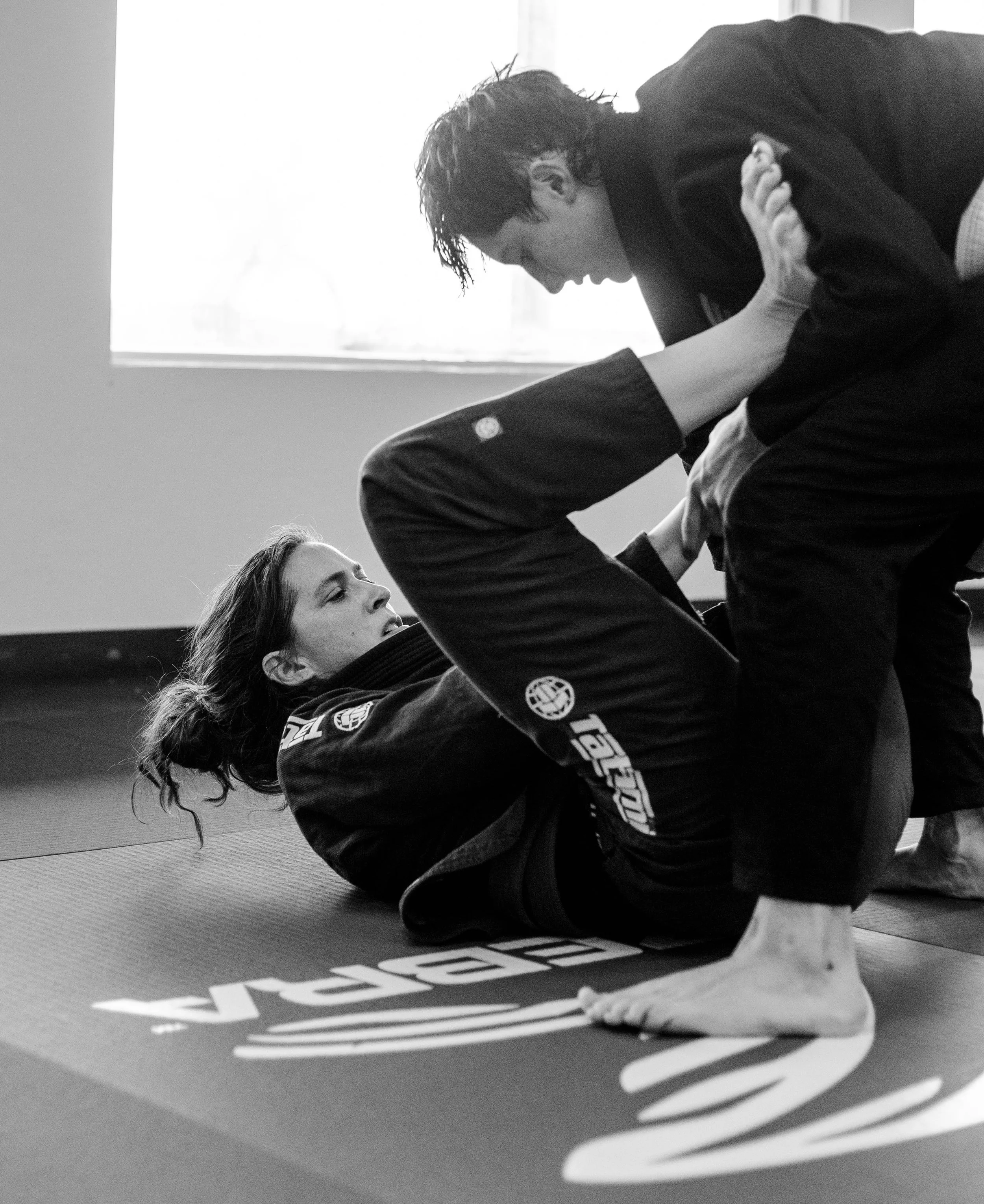 2 people practicing Brazilian Jiu Jitsu on a mat. A woman is on her back with her legs wrapped around the other person, who is leaning over her, holding her arm and neck. The setting appears to be a martial arts gym with a window in the background.