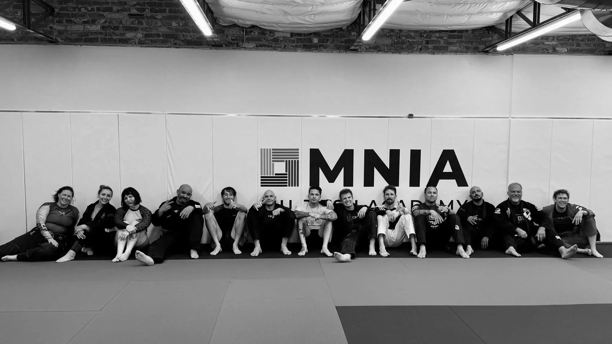 A group of people sitting on the floor at a martial arts gym, smiling and making gestures. They are in front of a wall with the logo 'Omnia' on it.