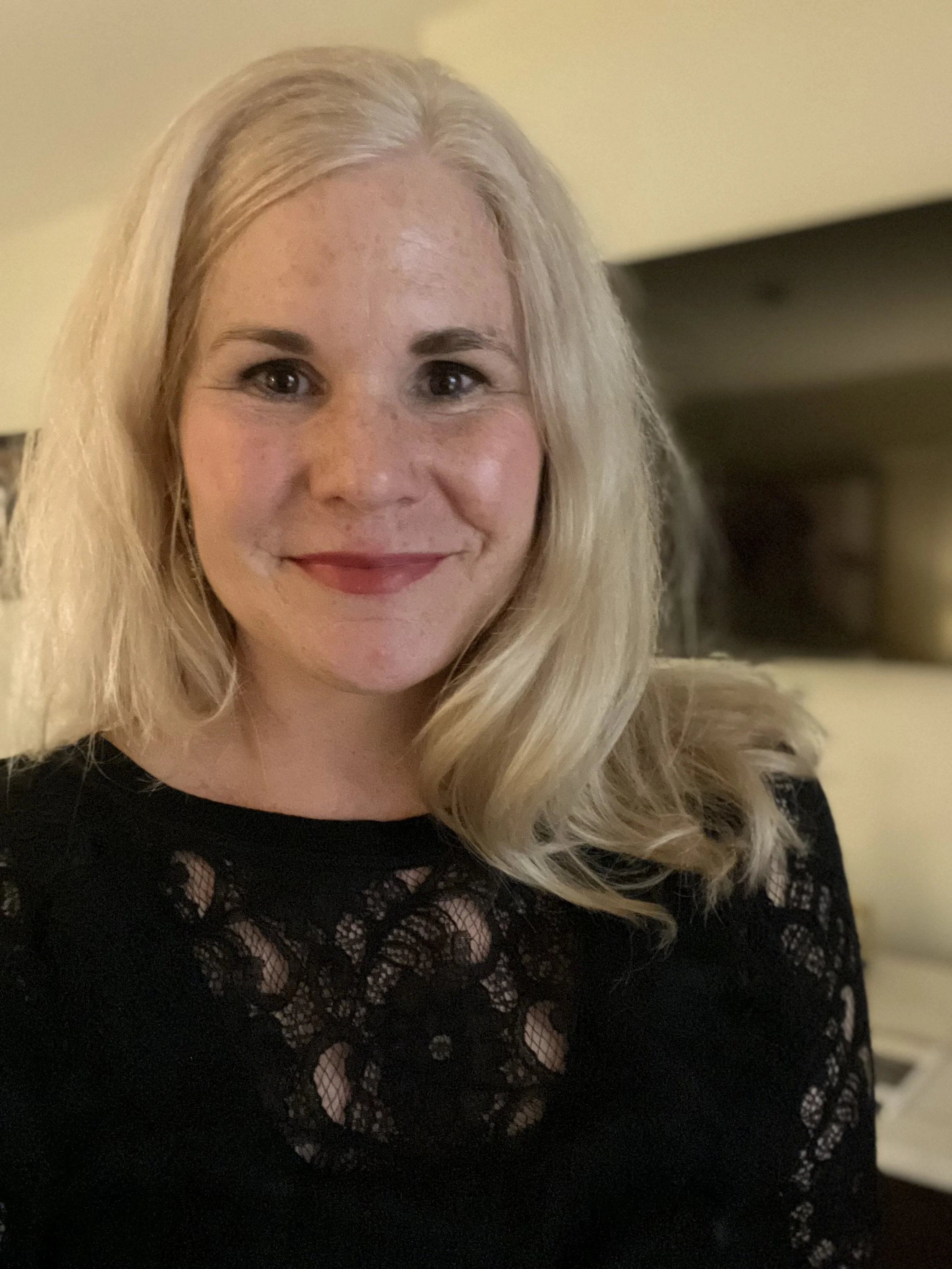 Portrait of a woman with blonde hair wearing a black lace top, smiling indoors with a blurred background.