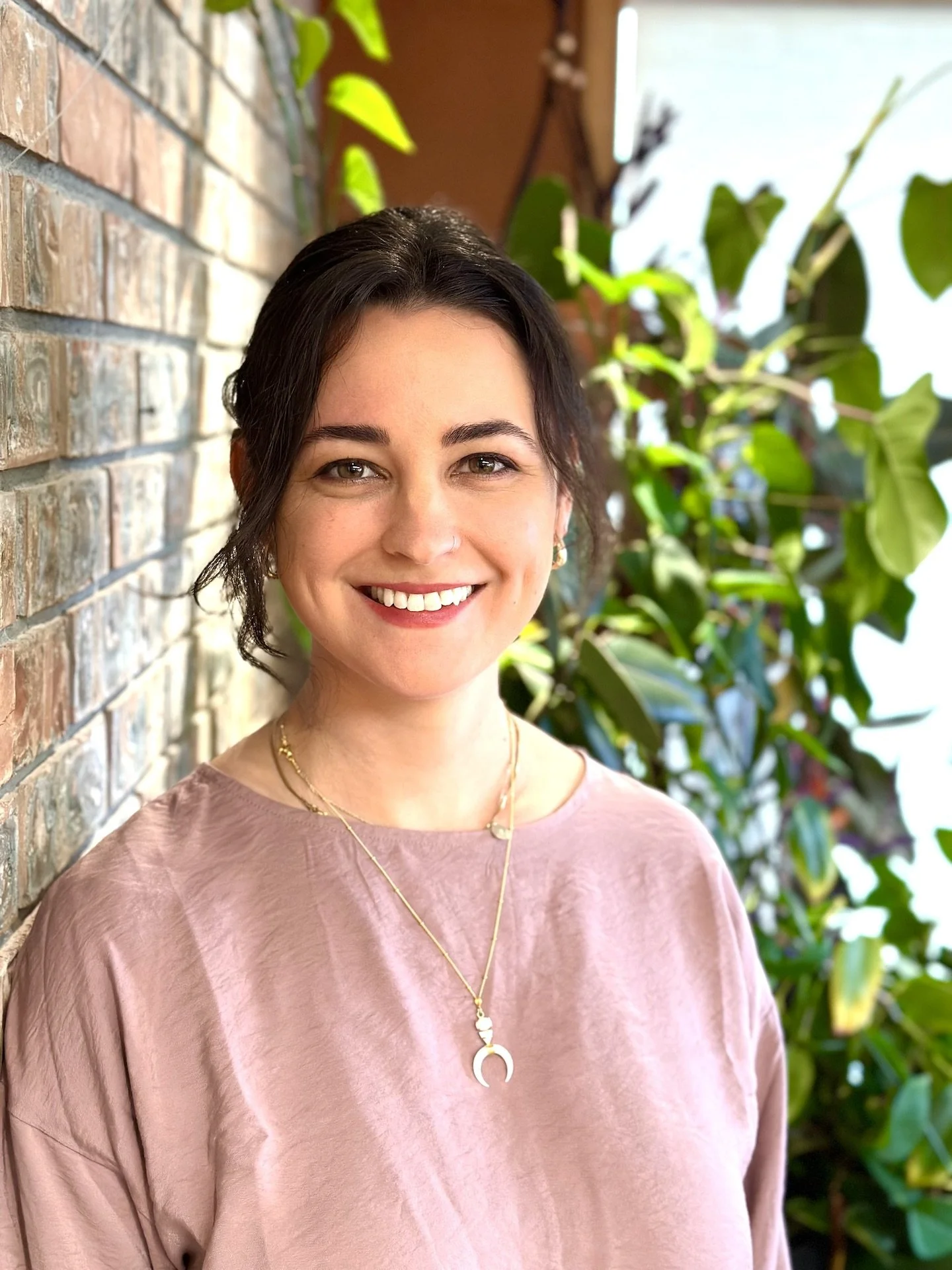 A young woman with dark brown hair, smiling, standing by a brick wall and green leafy plants.