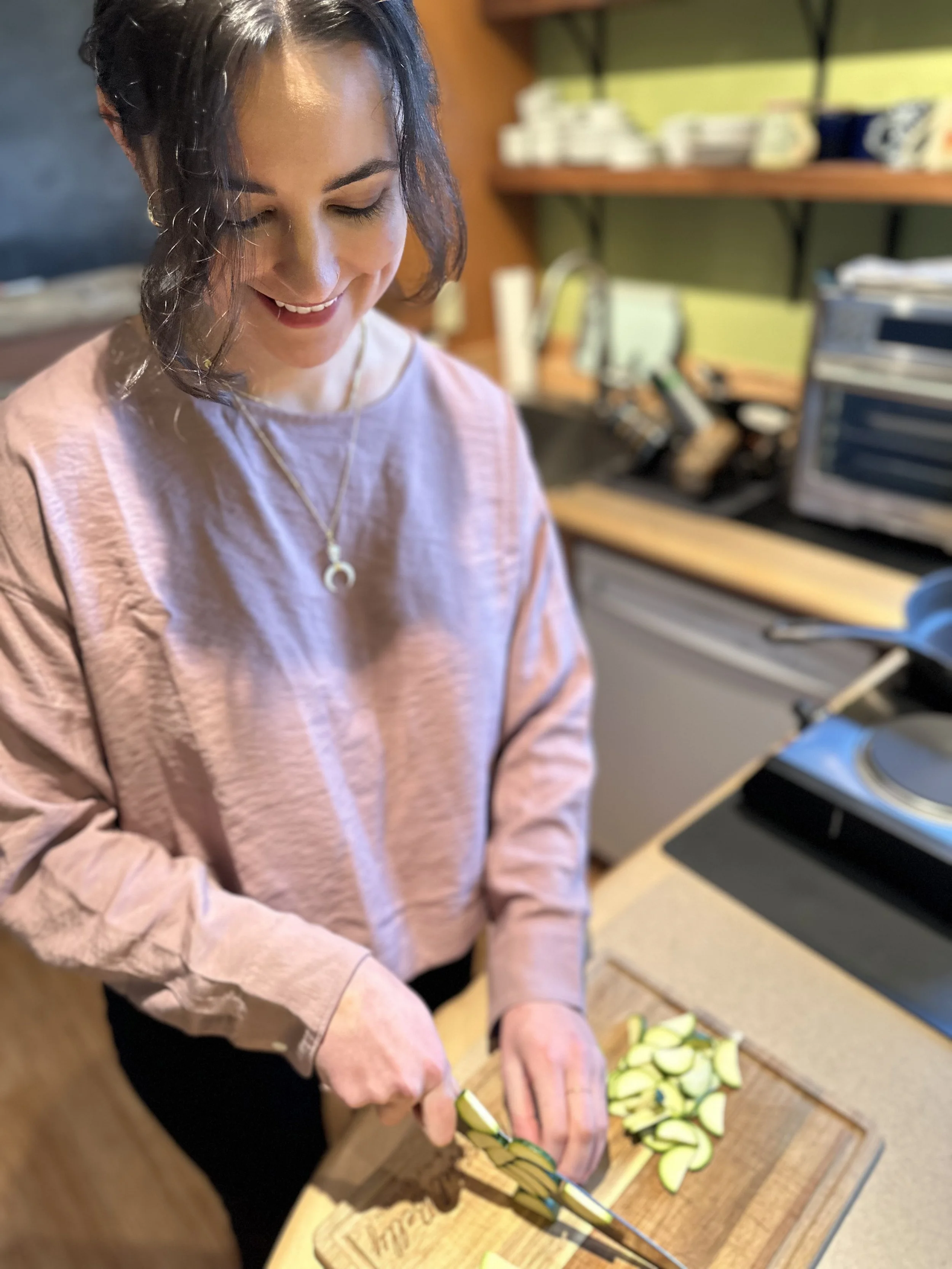 A woman with dark wavy hair, wearing a pink long-sleeve shirt and necklace, is slicing zucchini on a wooden cutting board in a kitchen.