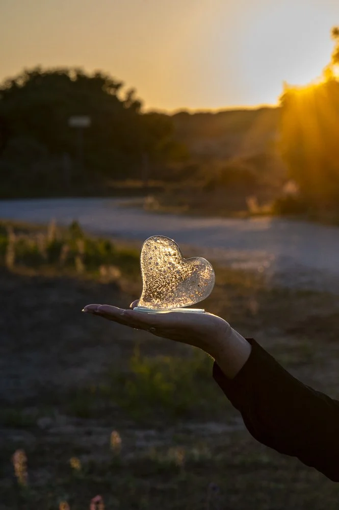 Een hand houdt een hartvormig stuk glas of ijs tegen de achtergrond van een zonsondergang met een rivier en bomen.
