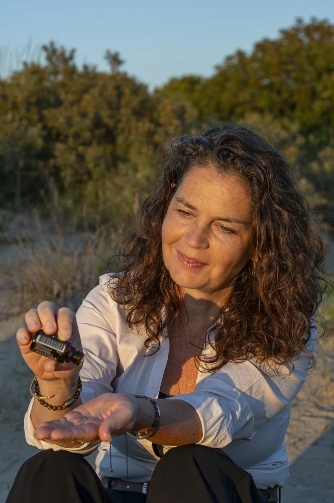 Vrouw die essentiële olie uit een flesje sprüht in haar hand in een natuurlijke buitenomgeving tijdens zonsondergang.