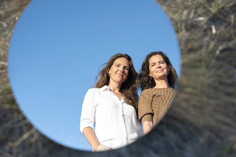Twee vrouwen kijken door een rond spiegel, met een blauwe lucht op de achtergrond.
