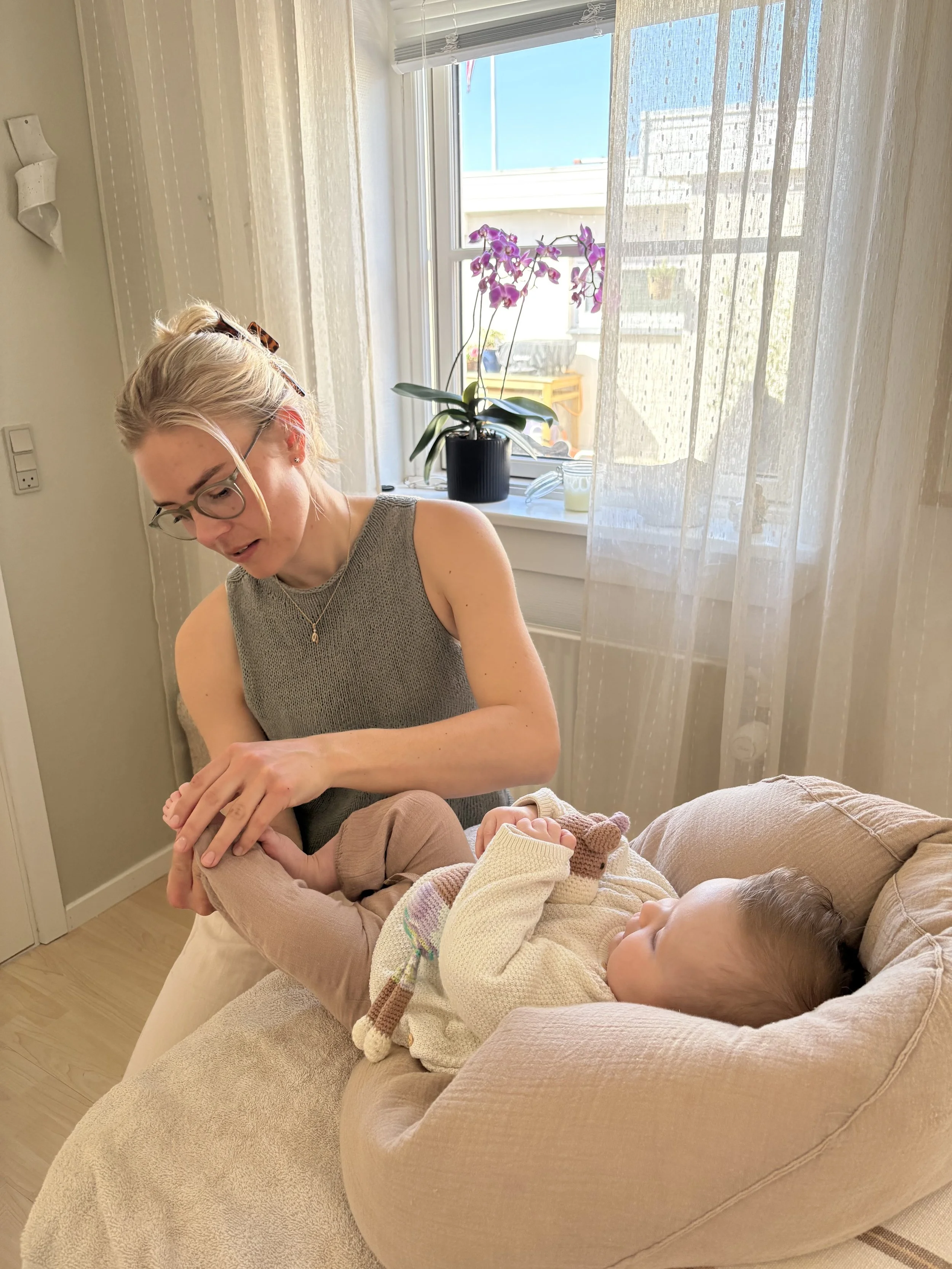 A woman with blonde hair, glasses, and a gray sleeveless top is sitting on a beige armchair, holding a child's foot and smiling. The child, lying on the armchair, is wearing a cream-colored sweater and has a toy sock on their hand. Behind them, there is a window with sheer curtains, a potted orchid on the windowsill, and a sunny outdoor scene.