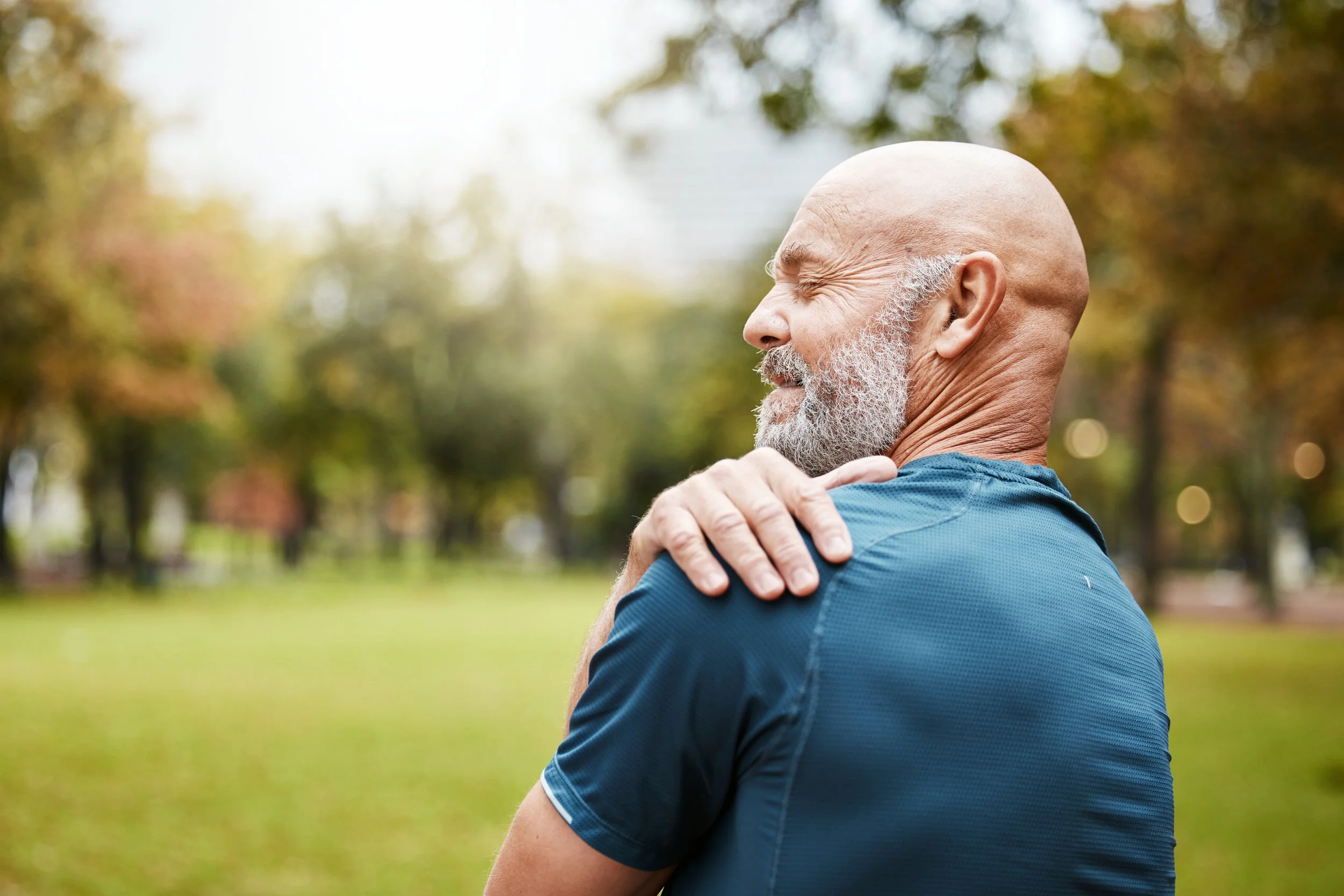 An elderly bald man with a white beard, smiling and stretching his shoulder in an outdoor park setting with green grass and trees in the background.