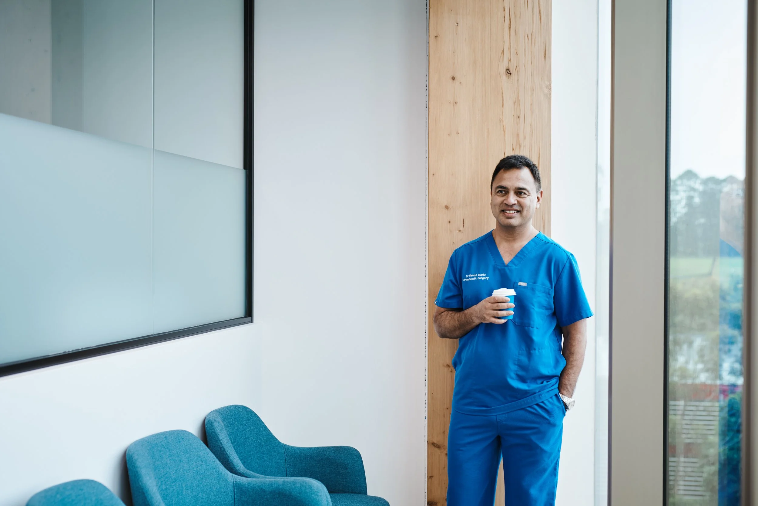 A male doctor or nurse wearing blue scrubs standing in a medical office, holding a coffee cup, smiling, with chairs and large windows in the background.
