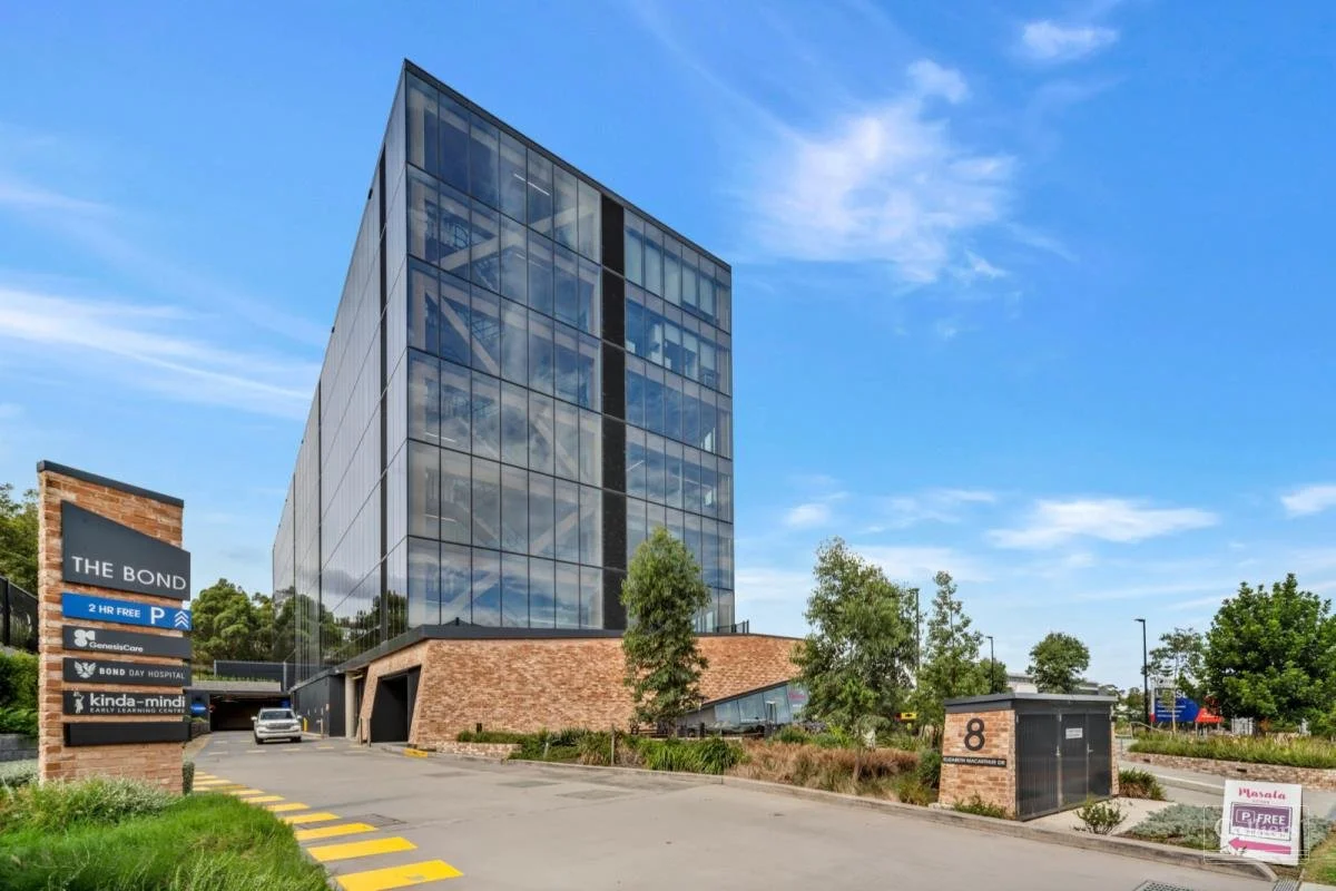 A modern office building with glass windows reflecting the blue sky and clouds, with a brick foundation and landscaped surroundings, including trees and a parking lot.