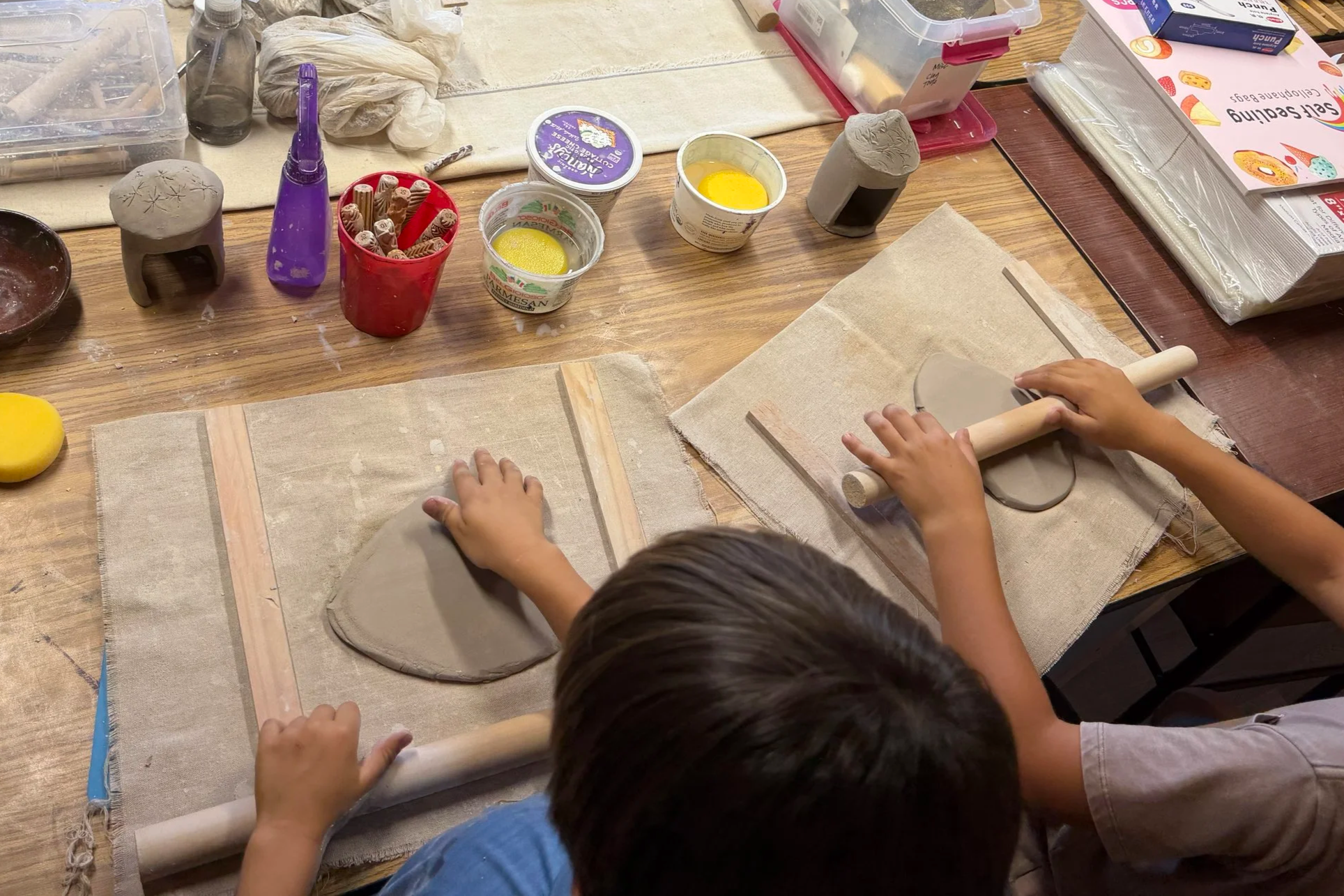 Person painting a colorful wooden craft piece with blue, yellow, and white paint, surrounded by paint jars, brushes, and art supplies on a wooden table.