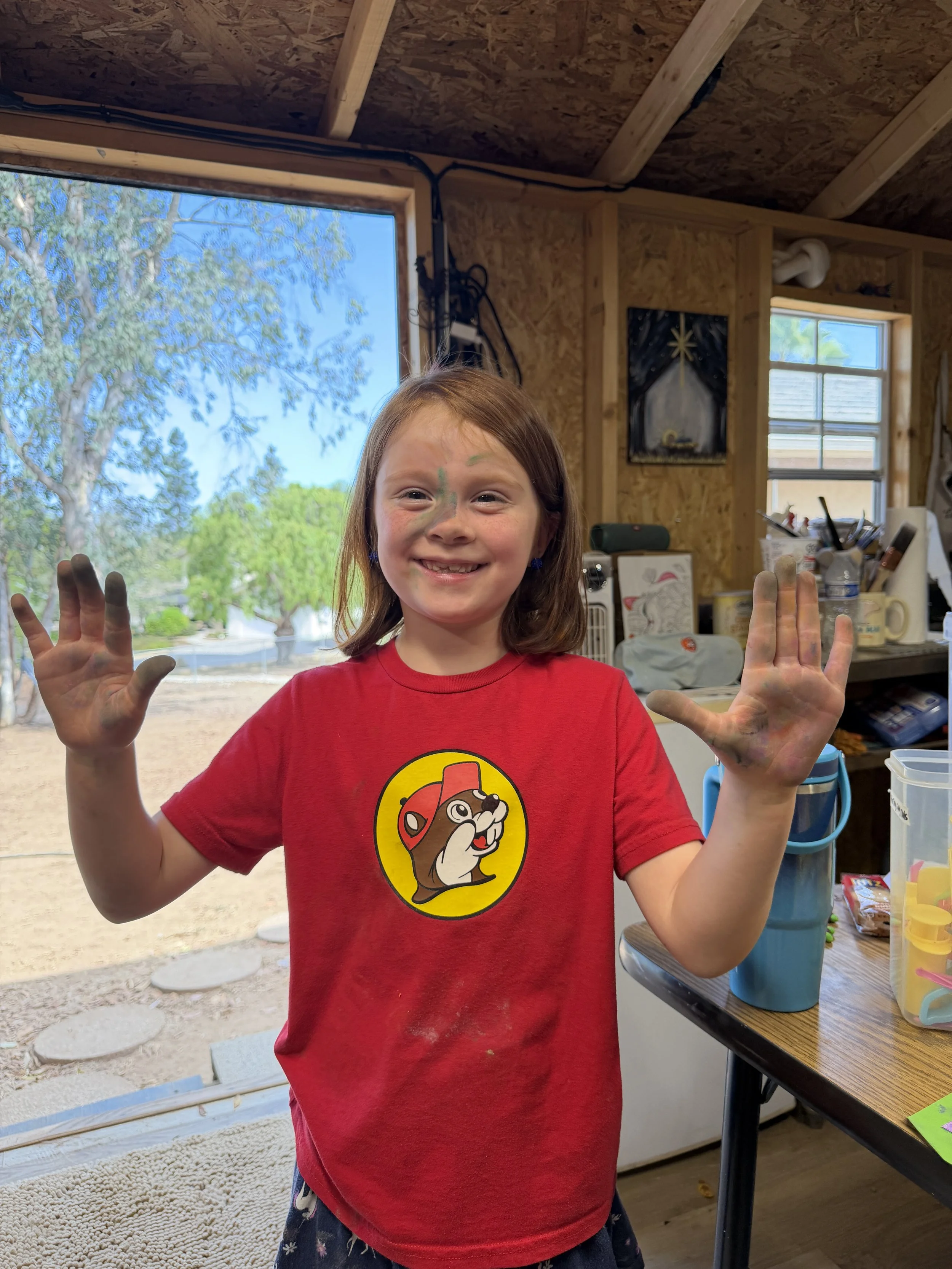A child's hand in a blue sleeve reaches over a messy table with colorful paint splatters, smears, and brushes, creating an abstract art scene with bottles of pink, orange, and purple paint surrounding the workspace.
