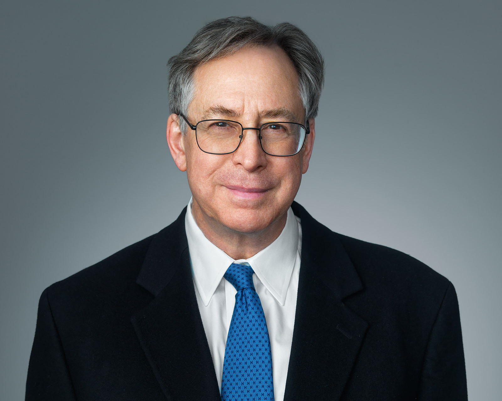 Portrait of a middle-aged man wearing glasses, a white shirt, a blue tie, and a black suit jacket against a plain gray background.