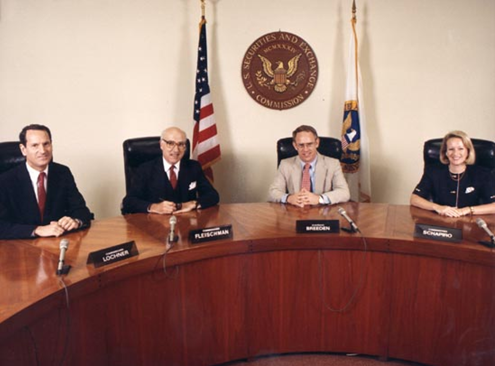 Four people sitting at a curved wooden table during a formal meeting, with an American flag and the SEC emblem behind them.