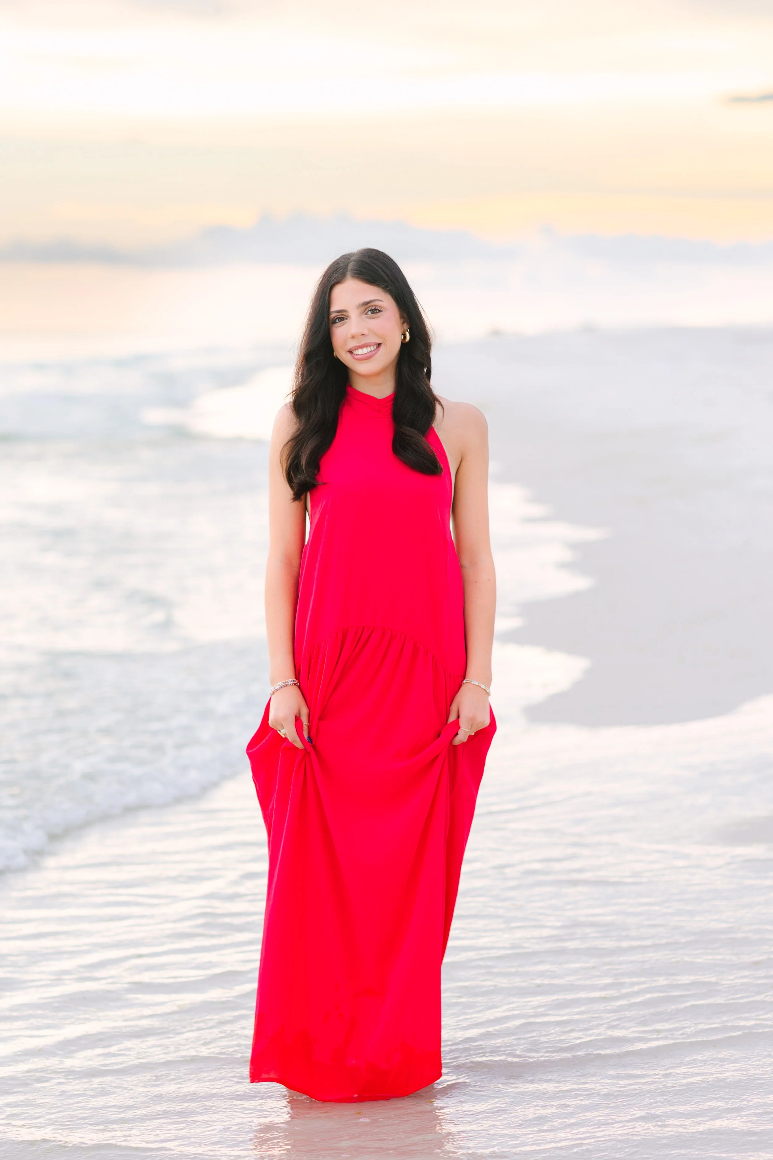 A young woman with long dark hair wearing a red dress standing in shallow water at the beach during sunset, smiling at the camera.