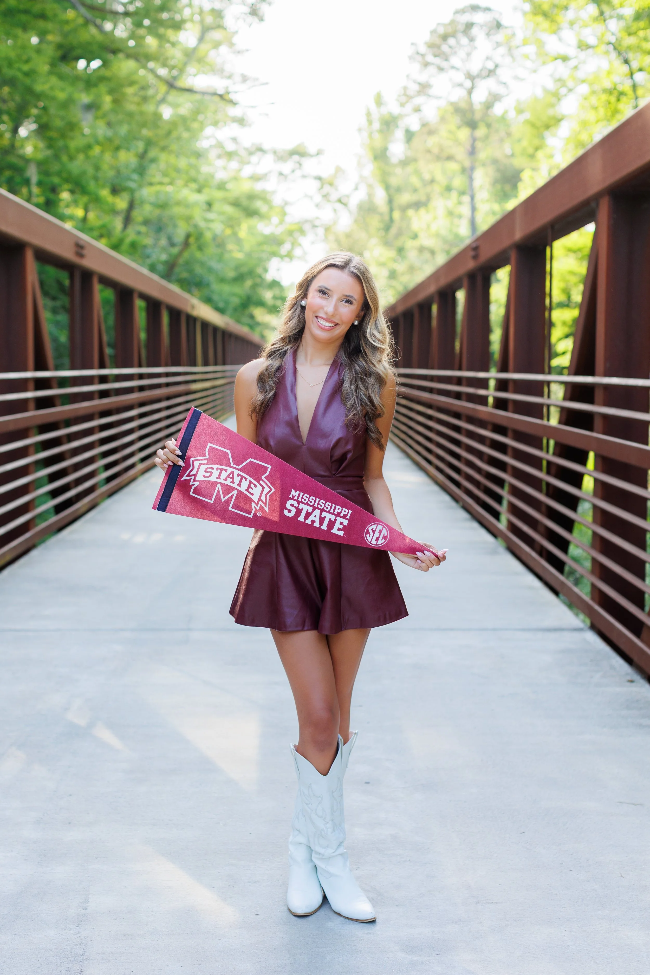 Young woman smiling and holding a Mississippi State pennant while standing on a bridge in a lush green, wooded park area.
