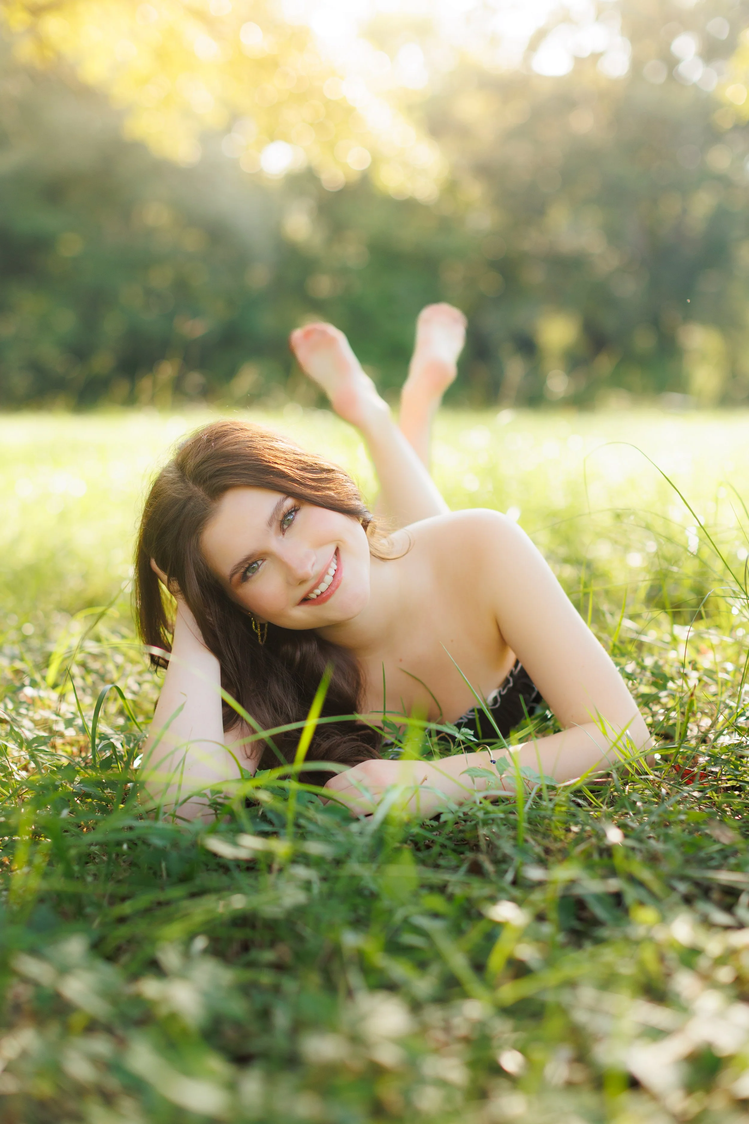 A young woman with long brown hair and blue eyes lying on a grassy field, smiling at the camera with trees and sunlight in the background.