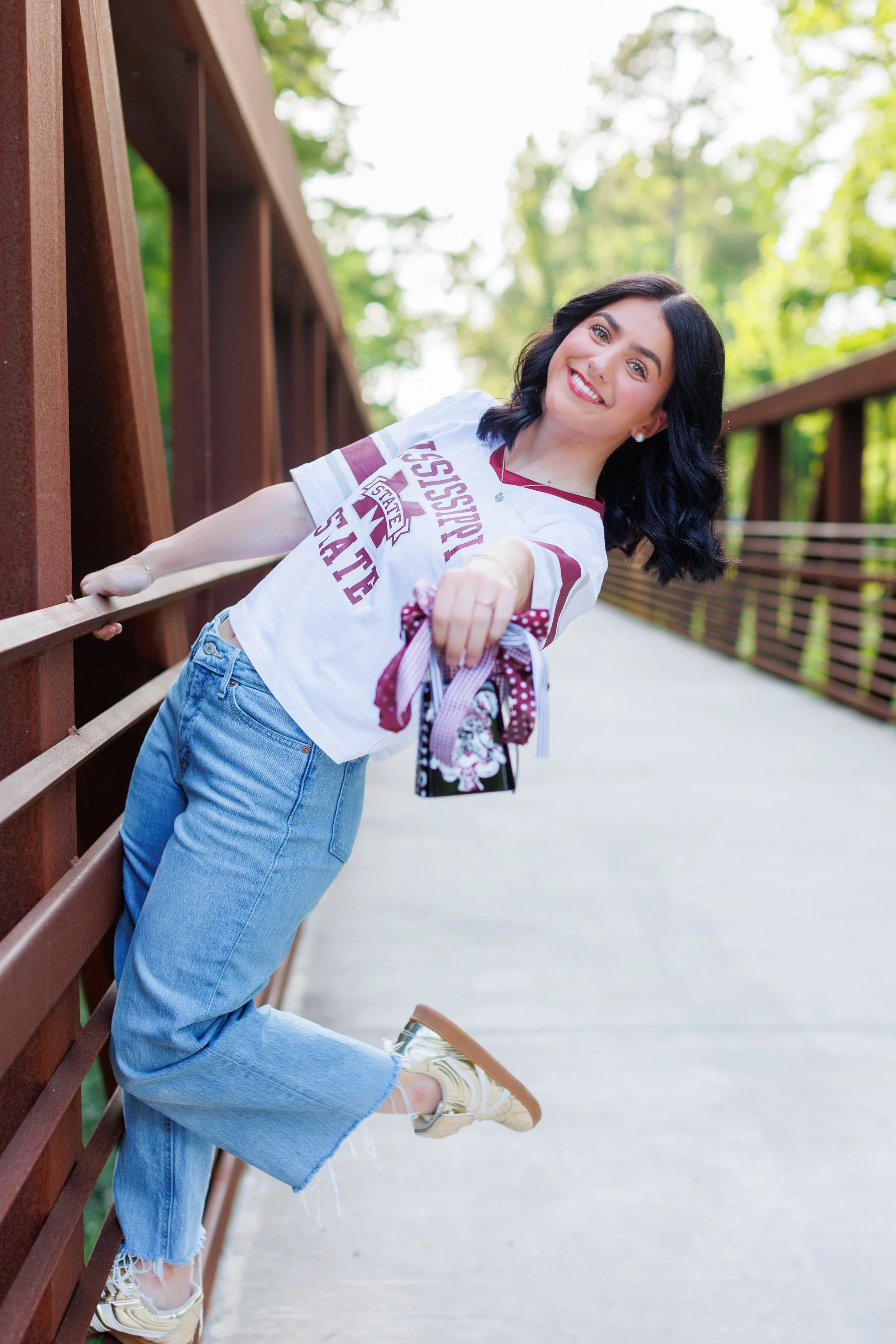 Smiling woman with black hair, wearing a white T-shirt with maroon lettering, light blue jeans, and sneakers, leaning against a wooden railing on a bridge in a park with green trees in the background.