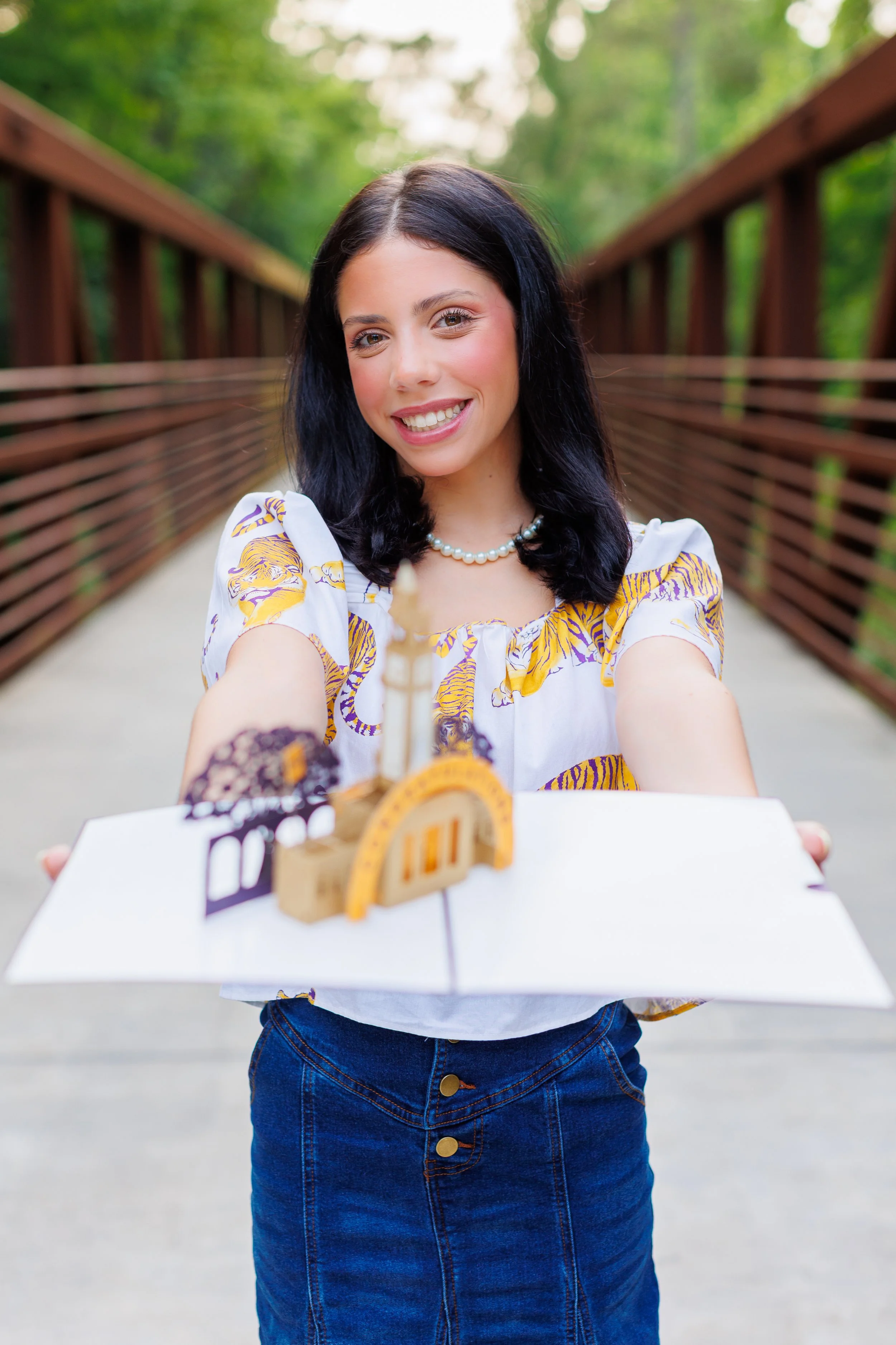 A woman with black hair taking a selfie with a smile, holding a small cardboard arch model on a tray in front of her. She's wearing a white and yellow patterned blouse, a pearl necklace, and a denim skirt. The background features a green, wooded outd
