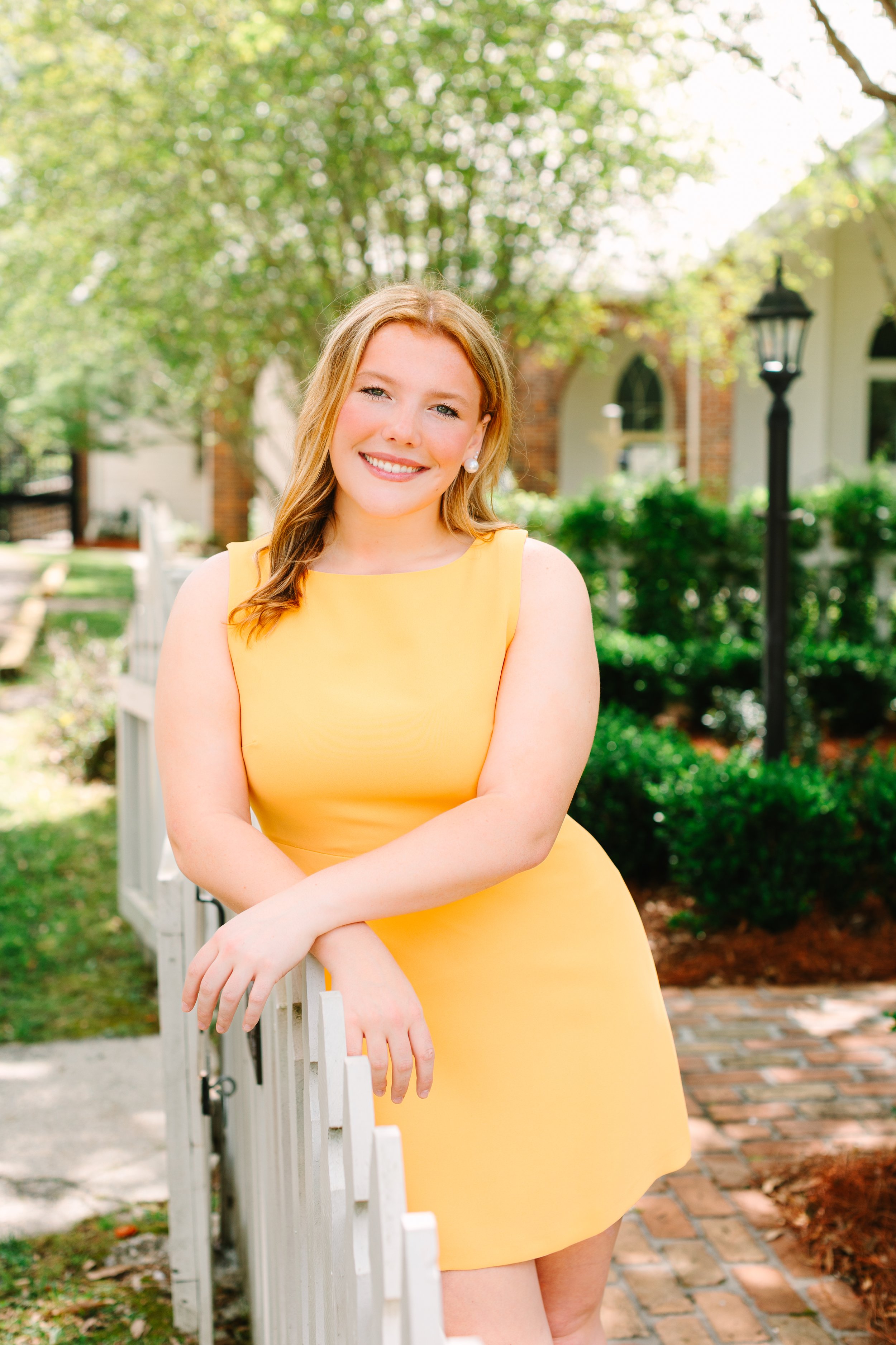 A woman with blonde hair in a yellow dress smiling outdoors, standing by a white fence with trees and a house in the background.