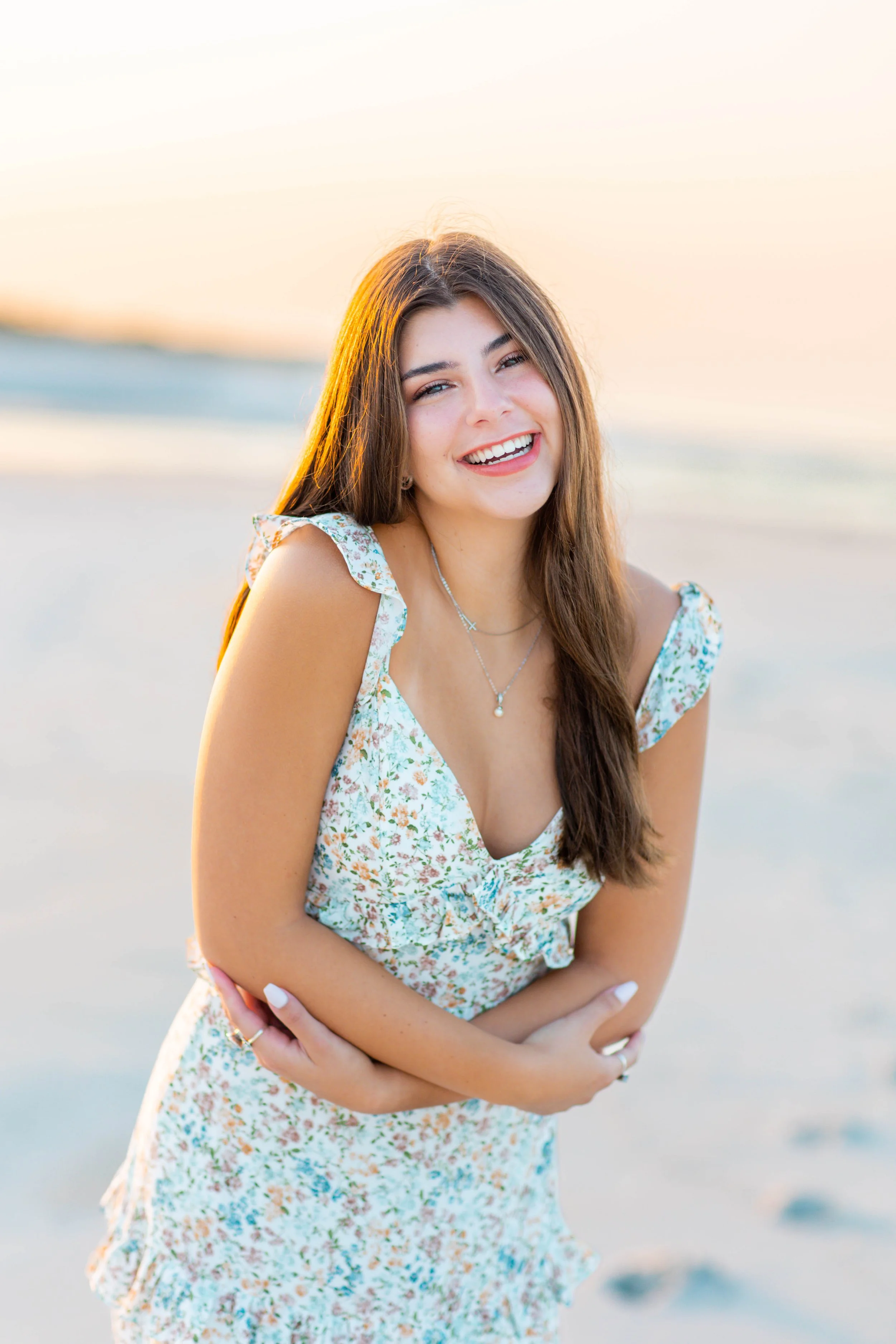 Smiling young woman with long brown hair wearing a floral dress on a beach at sunset.