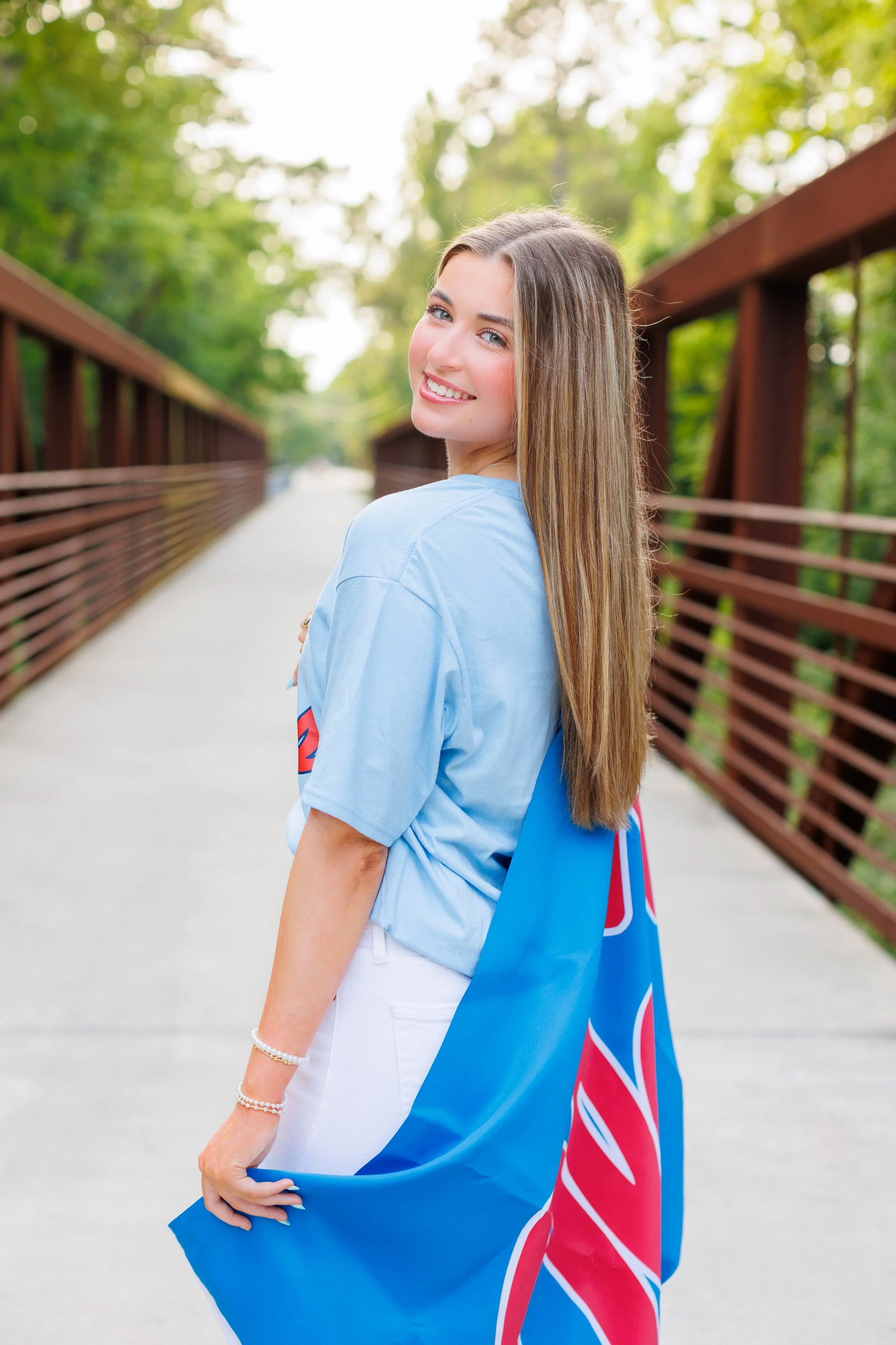 Smiling young woman with long blonde hair wearing a blue T-shirt and white pants, holding a Union Jack flag, standing on a bridge in a green park.