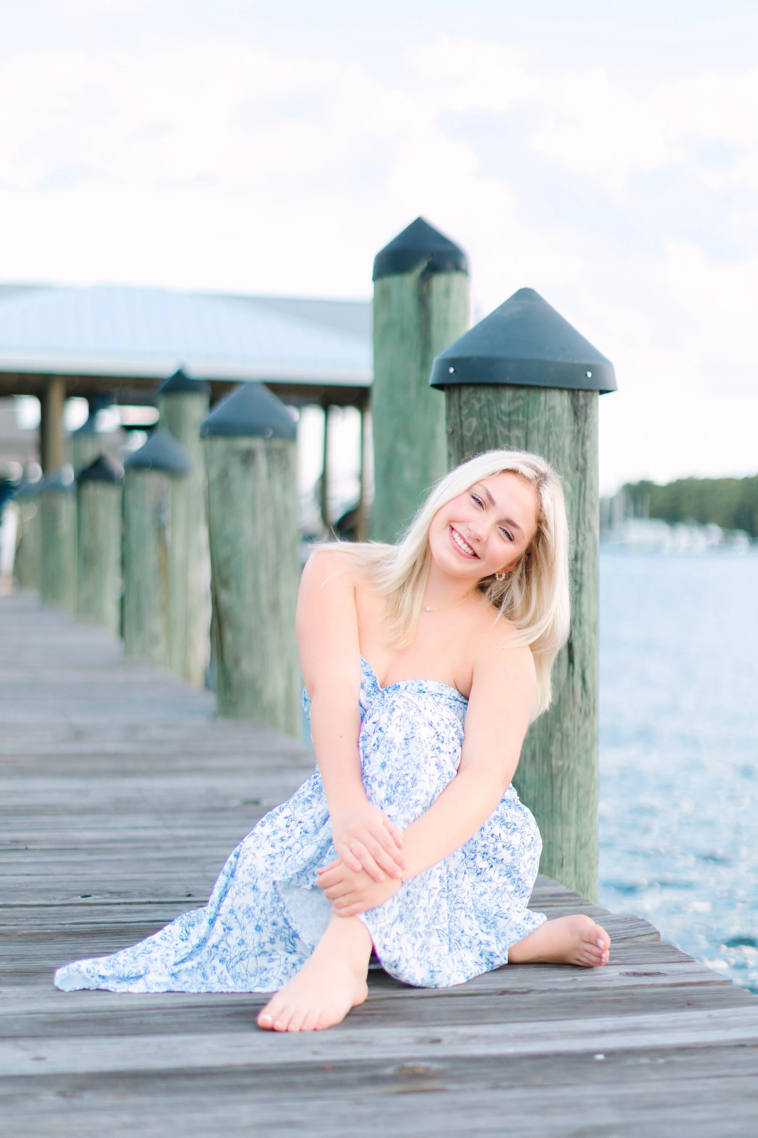 A young woman with blonde hair and a blue patterned dress sitting barefoot on a wooden dock by the water, smiling, with green wooden posts with black caps behind her.