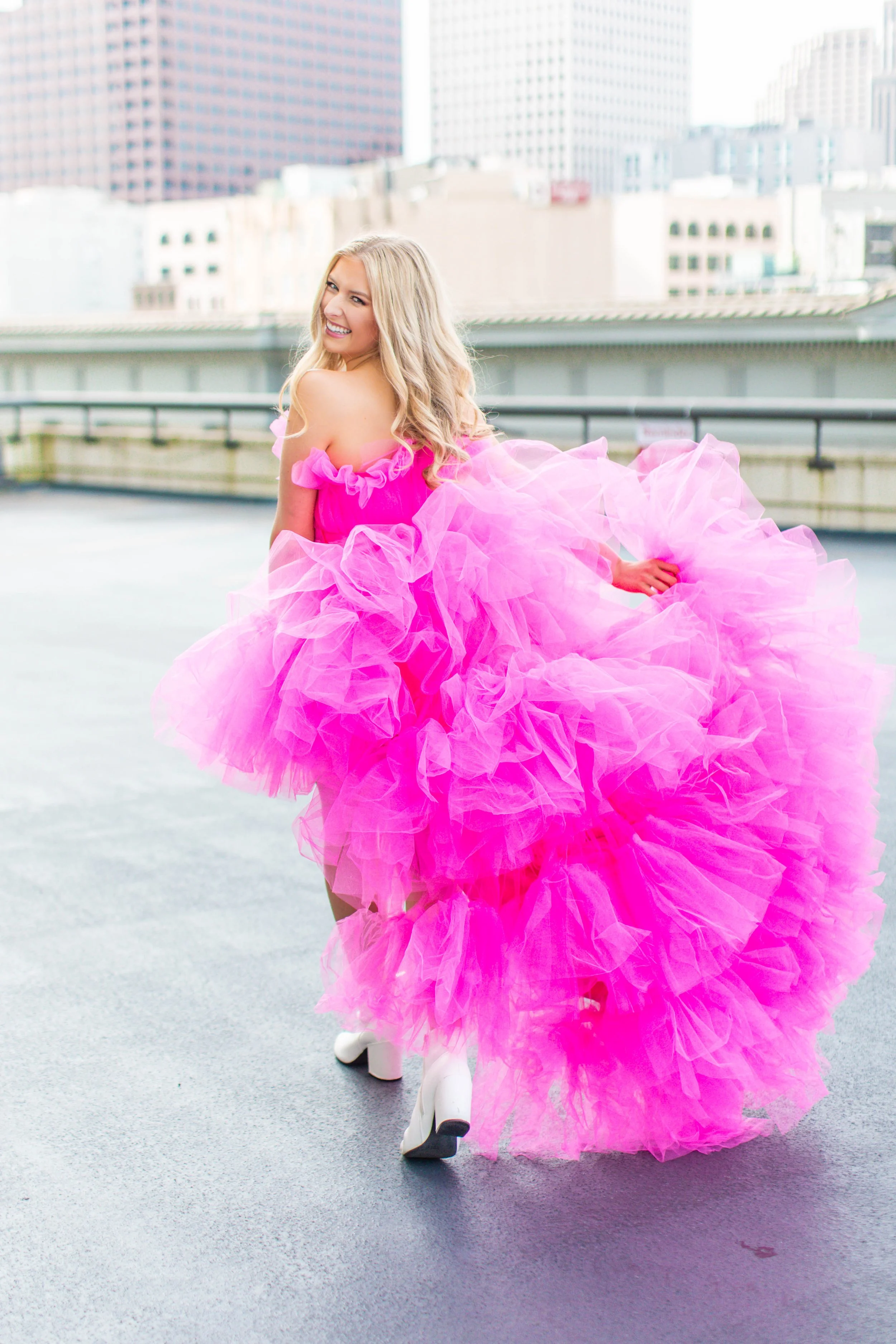 A woman with long blonde hair smiling and dressed in a bright pink, ruffled, voluminous gown standing outdoors in an urban setting with tall buildings in the background.