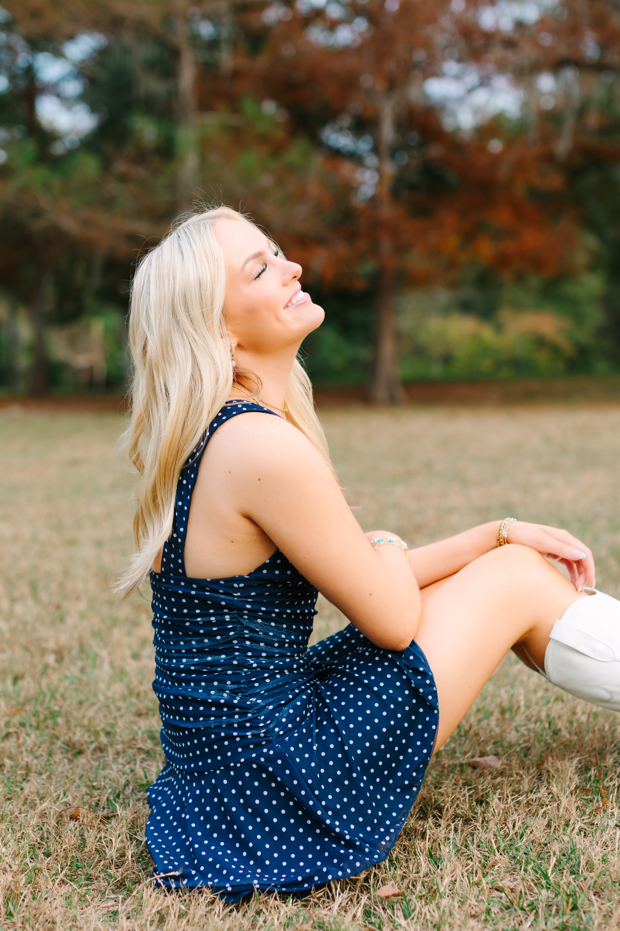 A young woman with long blonde hair, smiling and relaxing outdoors on a fall day. She is wearing a navy blue polka dot dress and white knee-high boots, sitting on the grass with trees showing autumn leaves in the background.