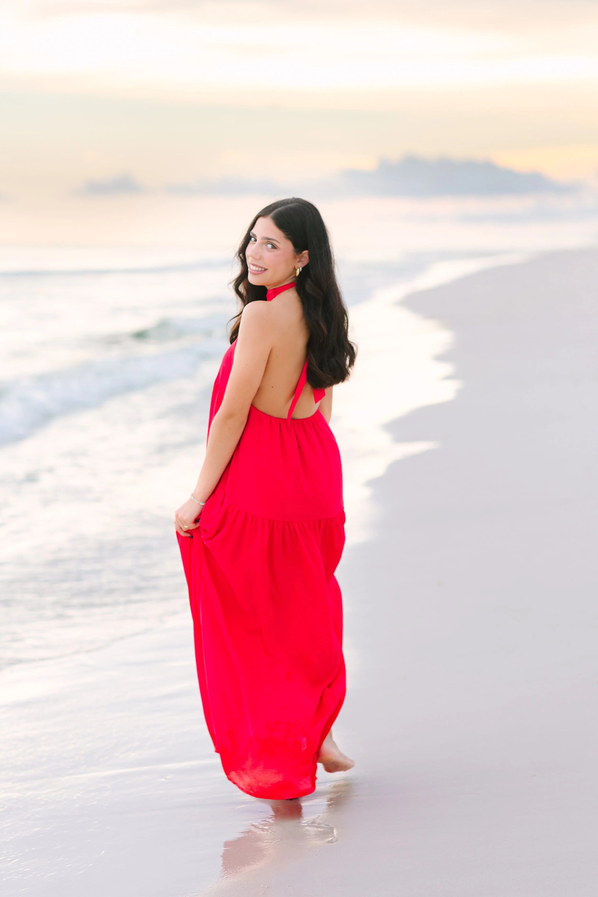 A woman with long dark hair wearing a bright pink halter dress is standing on a sandy beach, with her feet in the shallow water, during sunset or sunrise.