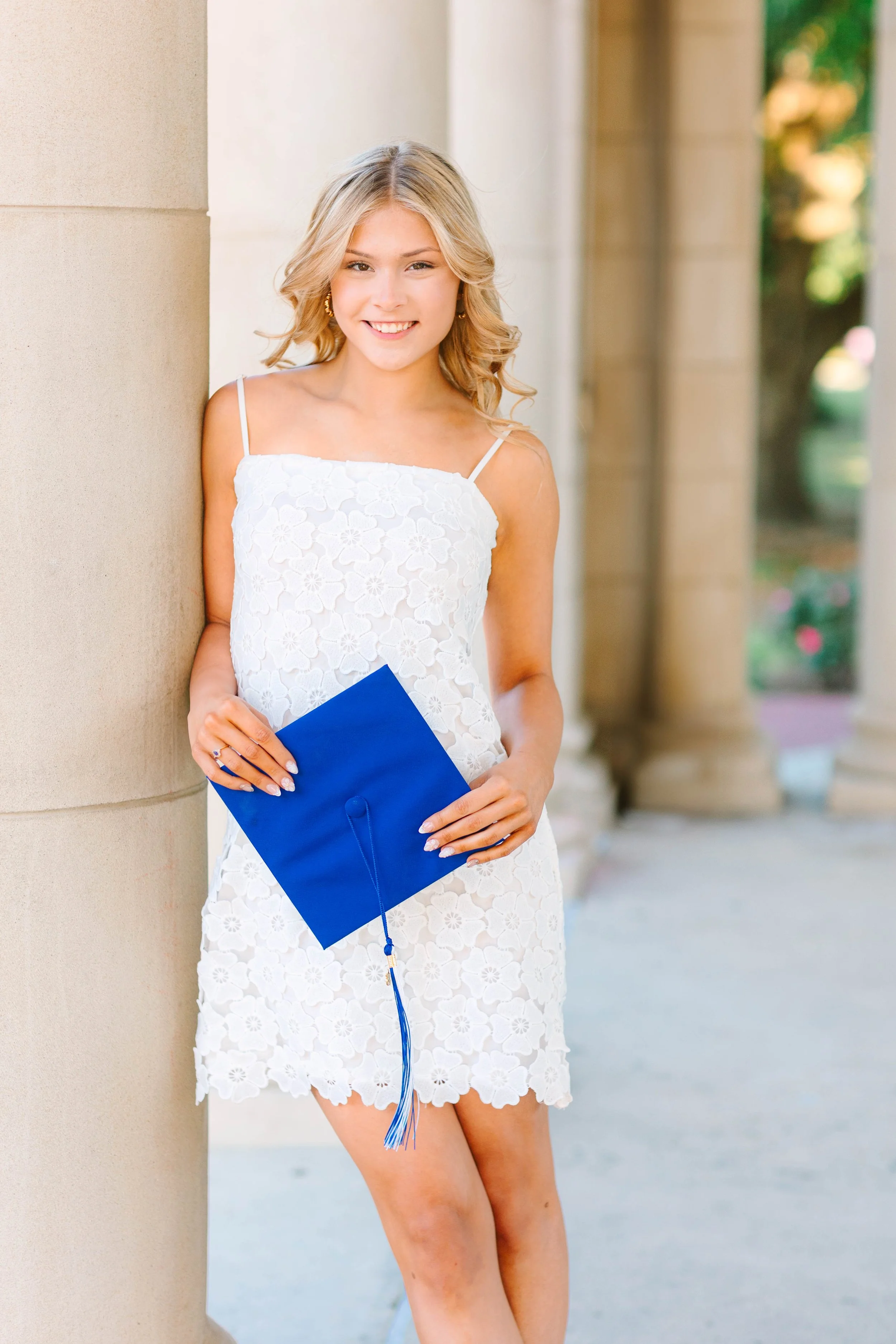 Young woman in a white lace dress holding a blue diploma, standing outside leaning against a beige column, smiling.
