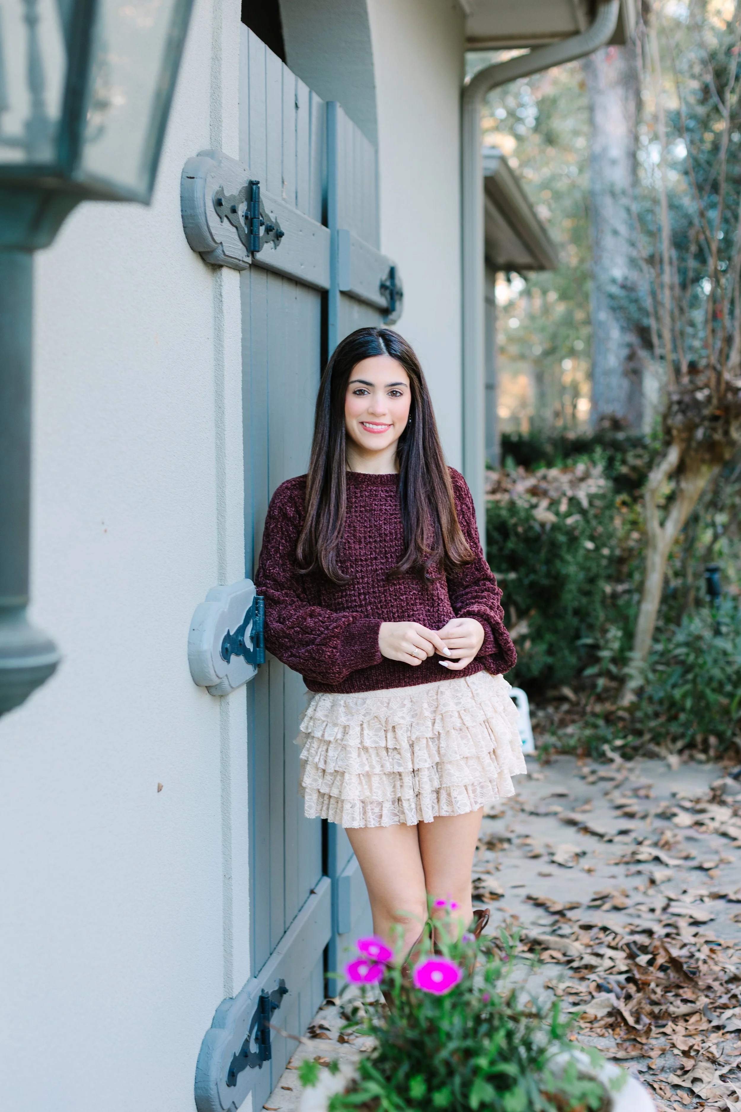 Young woman with long dark hair wearing a burgundy sweater and a tan ruffled skirt, standing outdoors near a house with light-colored walls, lots of trees and fallen leaves in the background, and pink flowers in the foreground.