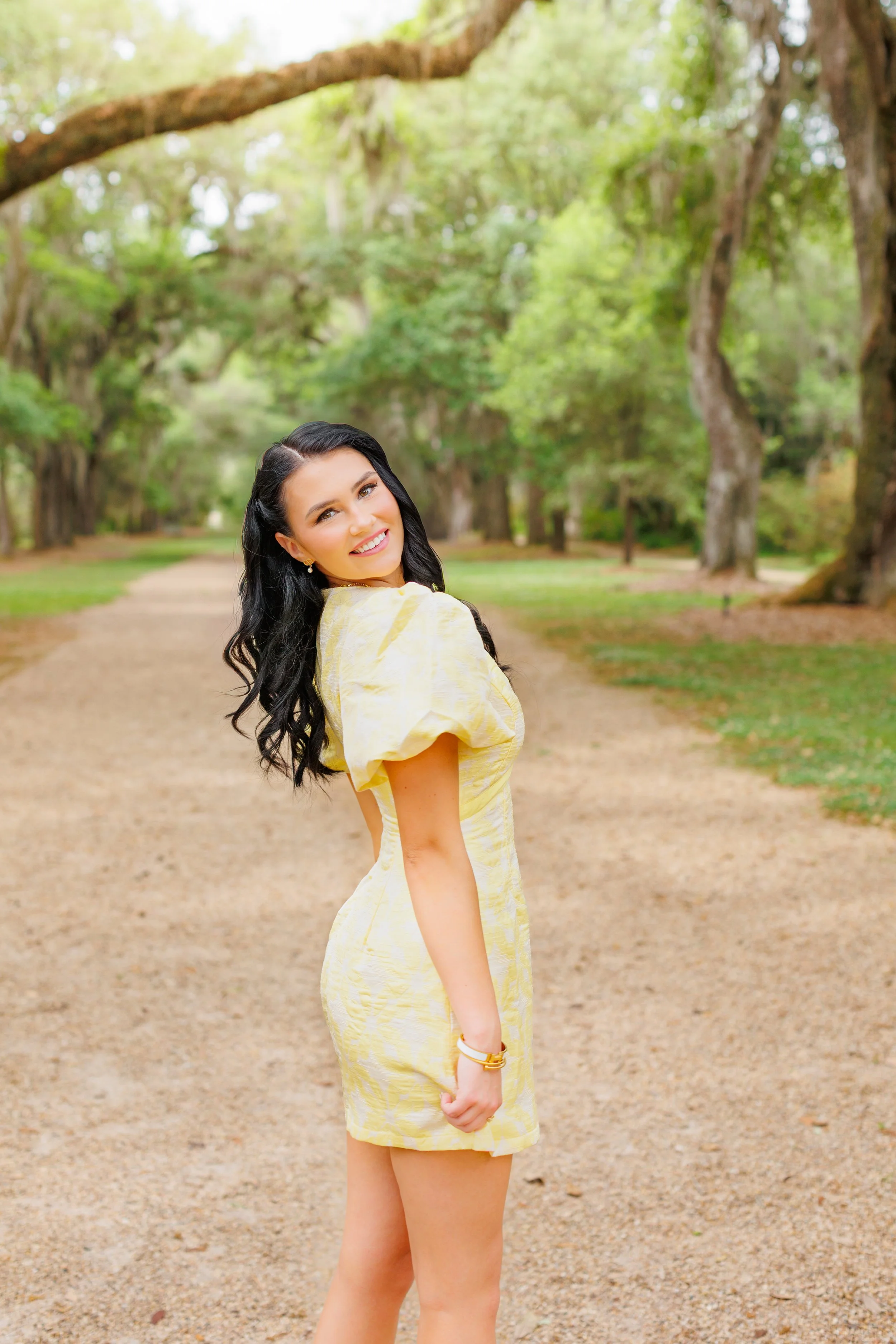 A woman with long black hair, wearing a yellow dress, smiling while standing on a dirt path in a lush green park with tall trees.