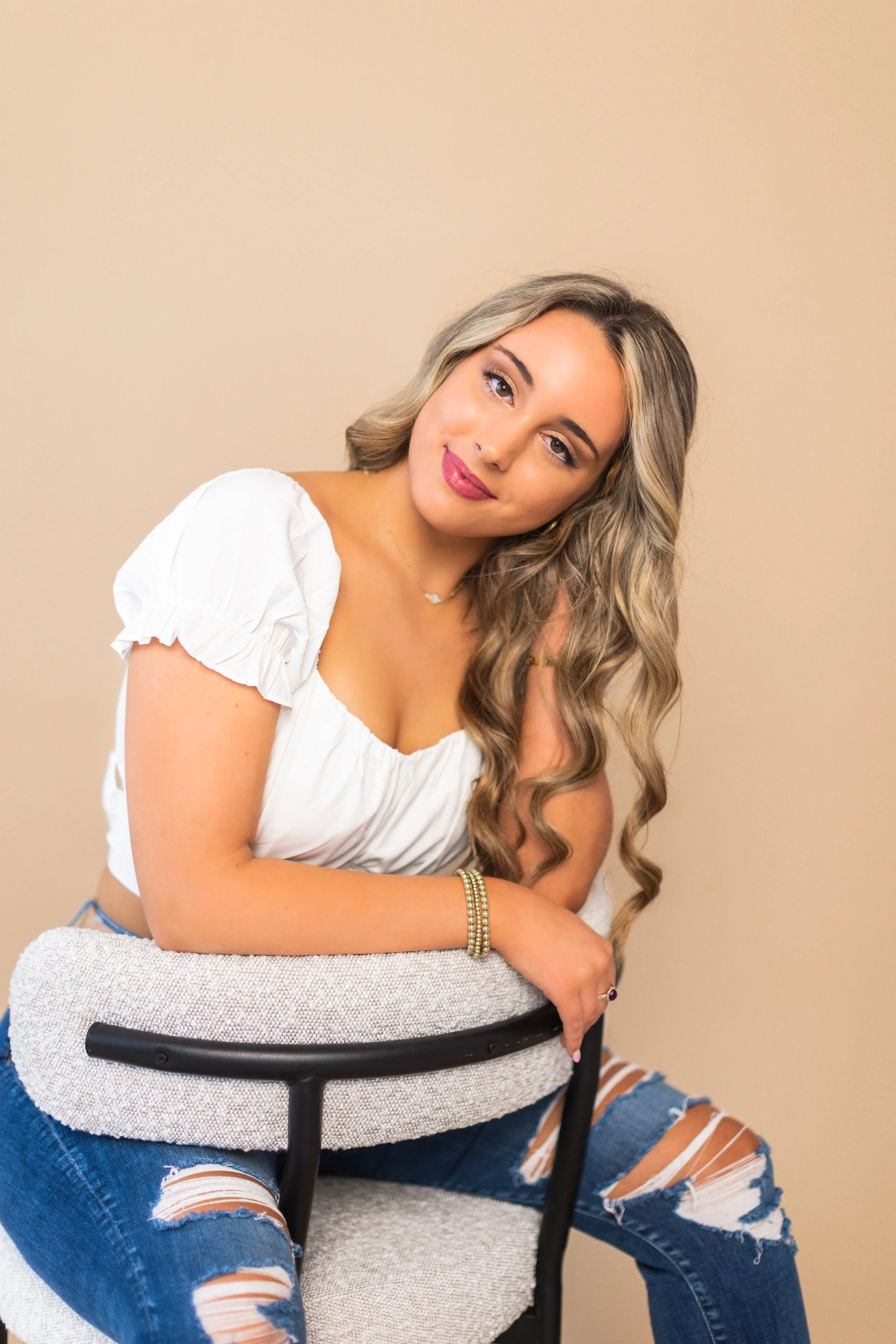 A woman with long wavy blonde hair, wearing a white puff-sleeve top, ripped blue jeans, and jewelry, sitting on an upholstered chair with her arm resting on her knee, against a plain beige background.