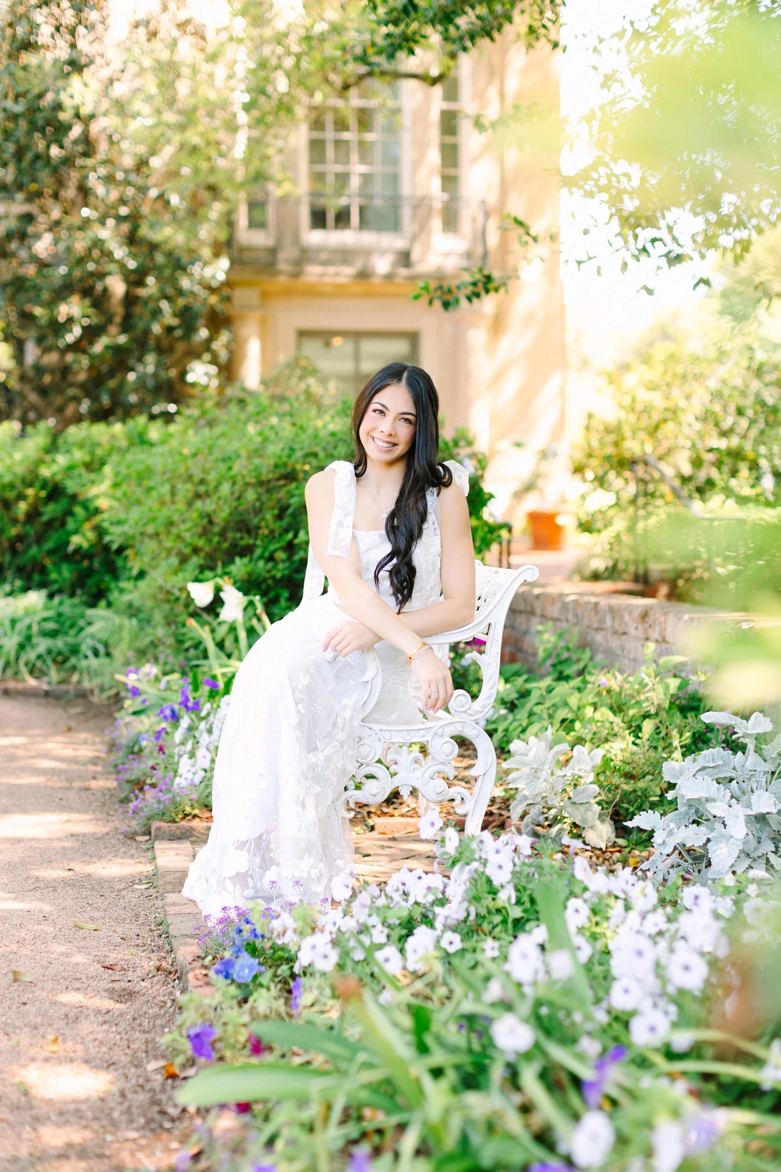 A young woman in a white dress sitting on a white ornate garden bench, smiling, surrounded by colorful flowers and greenery, with a house in the background.