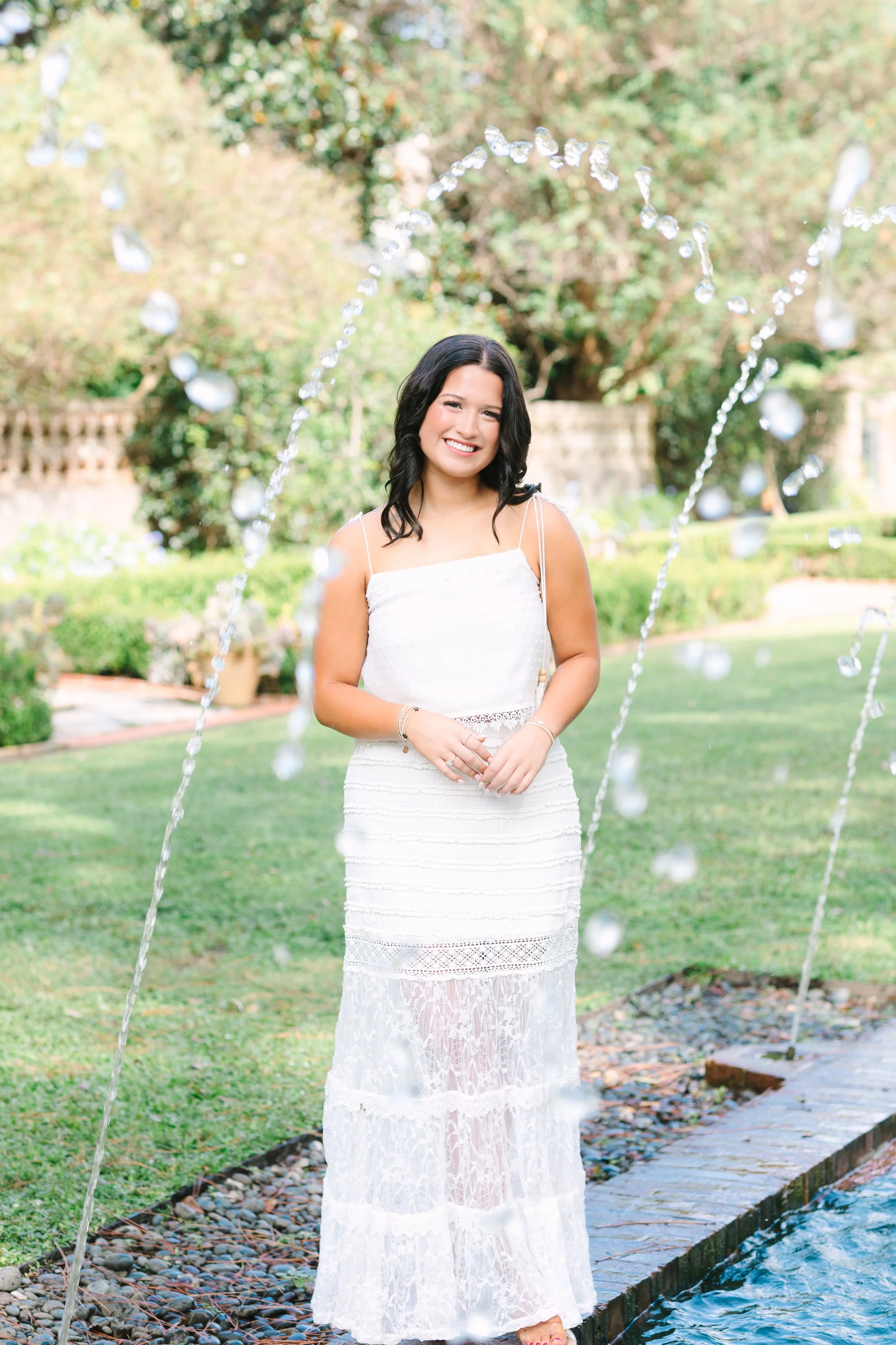 A woman with dark hair smiling in a white dress stands near a fountain in a garden with trees and grass.