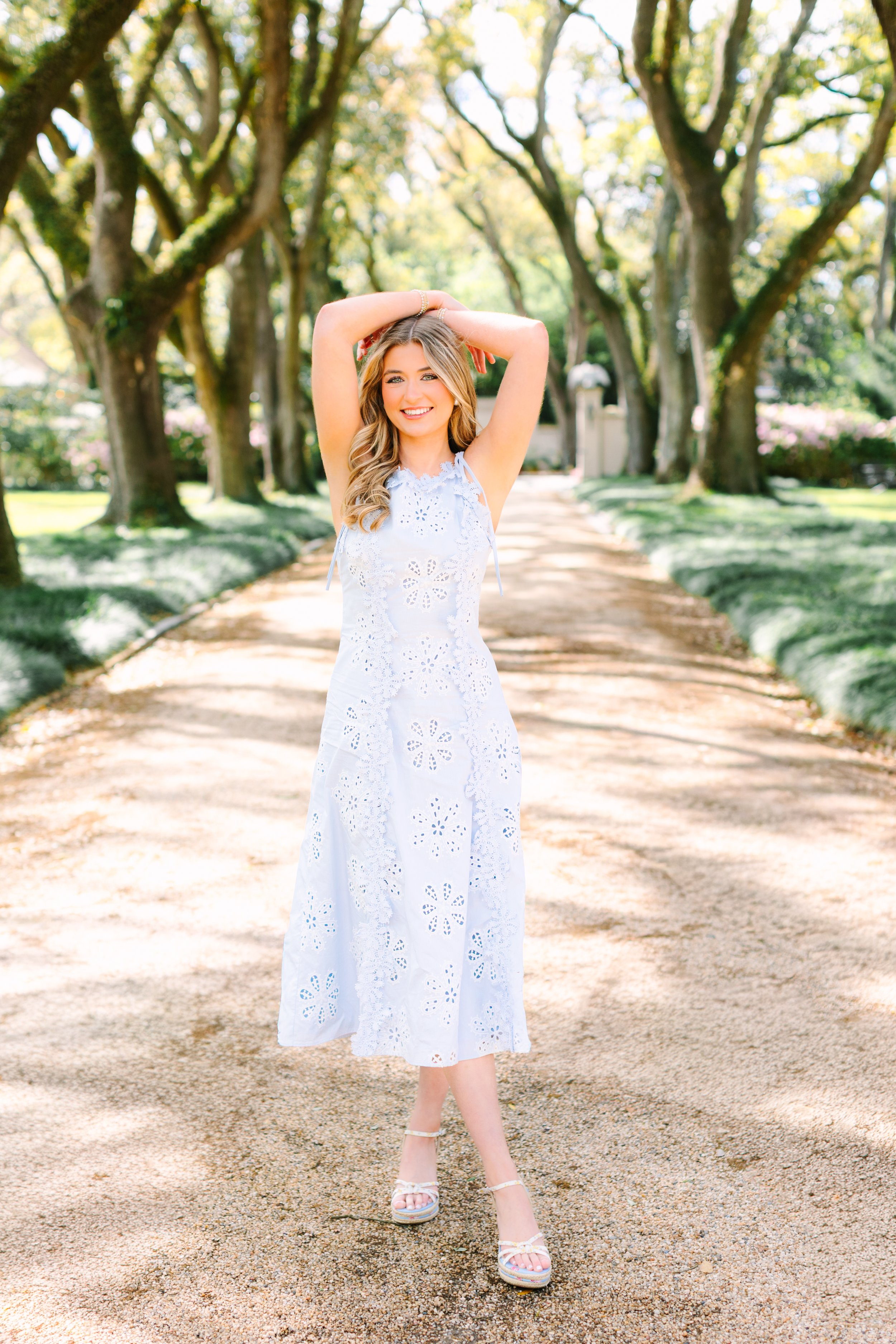 Young woman in white eyelet dress and platform sandals standing on a gravel path lined with trees, smiling with arms raised behind her head.