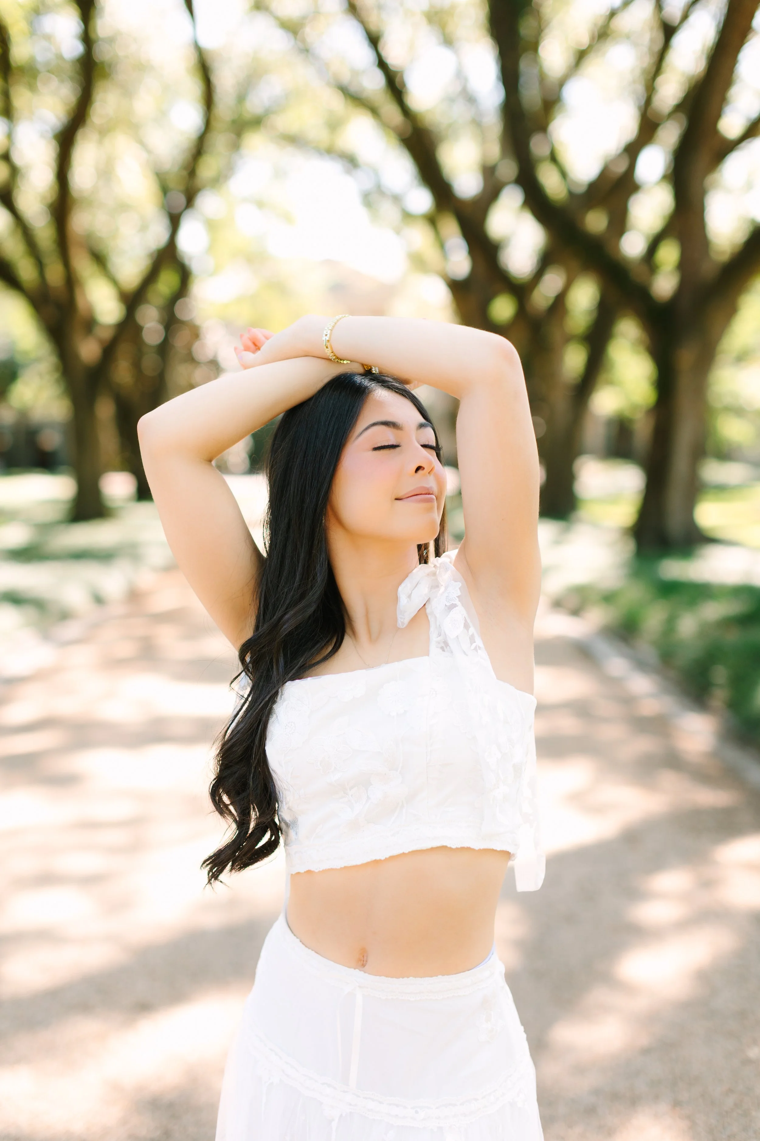 A woman with long black hair and closed eyes standing outdoors with her arms raised above her head, wearing a white lace top and a bracelet, surrounded by trees with green leaves and blurred sunlight.