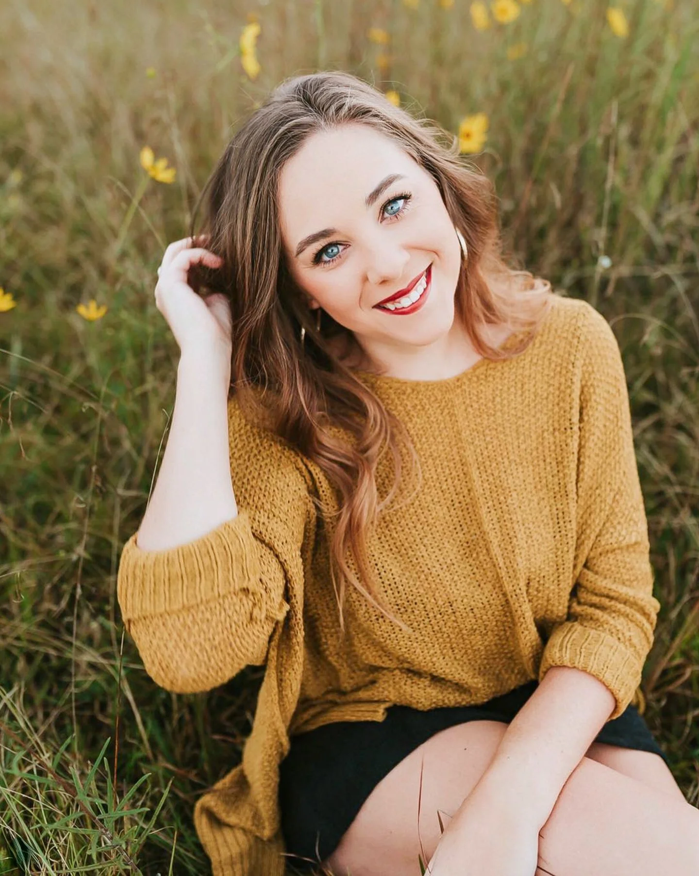 A woman with blue eyes, red lipstick, and brown wavy hair sitting in a field of tall grass and yellow flowers, smiling and touching her hair.