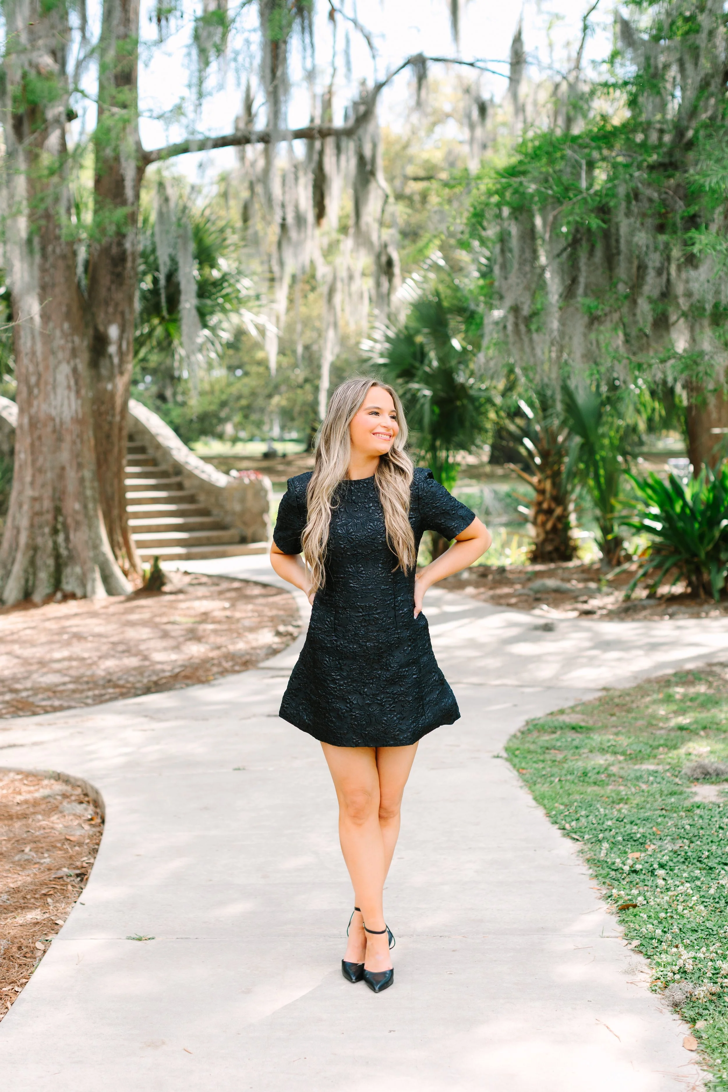 A woman with long blonde hair wearing a short black lace dress and black heels standing on a sidewalk in a park with trees and stairs in the background.