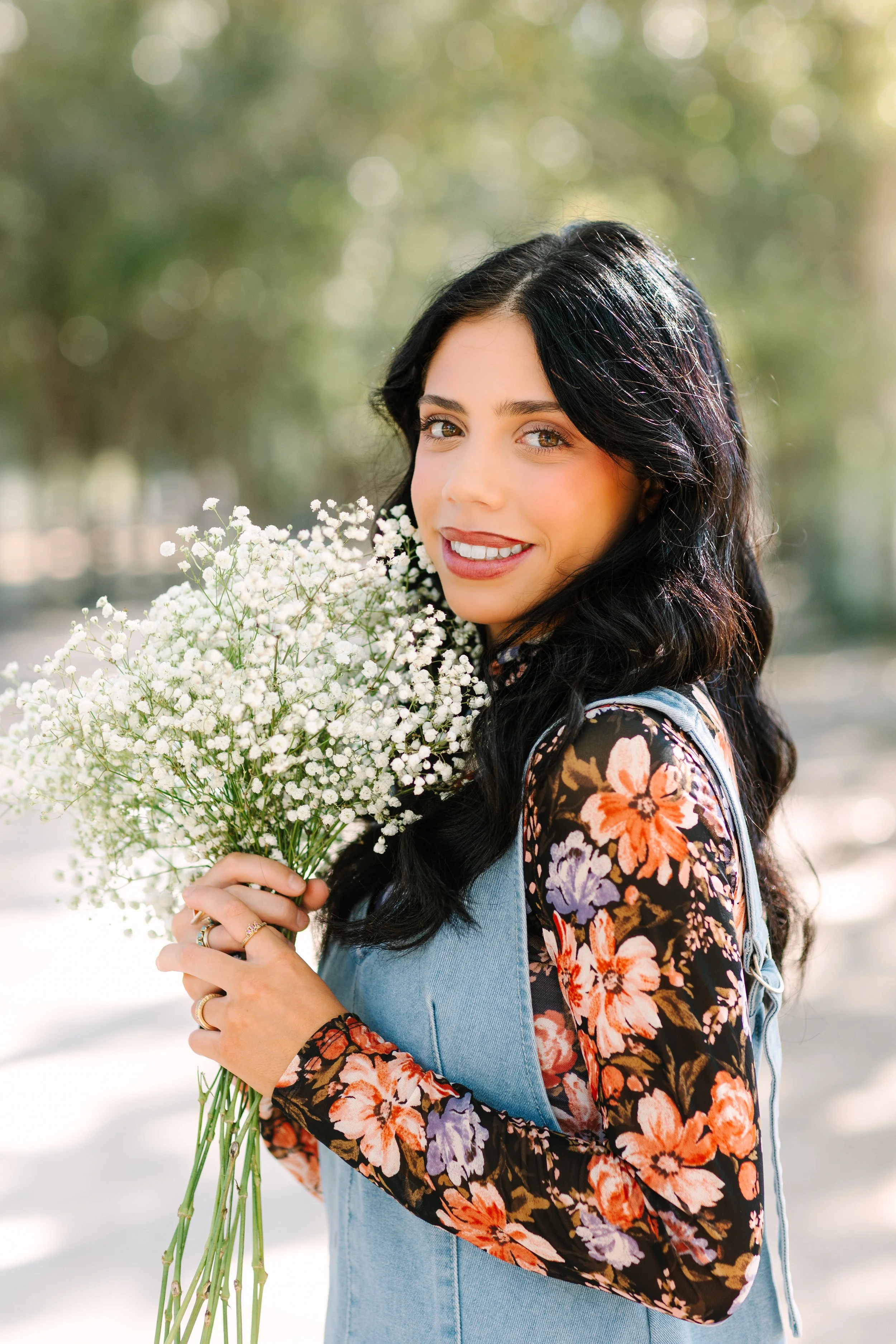 A woman with long dark hair holds a bouquet of small white flowers outdoors in a sunny setting with green blurred background.