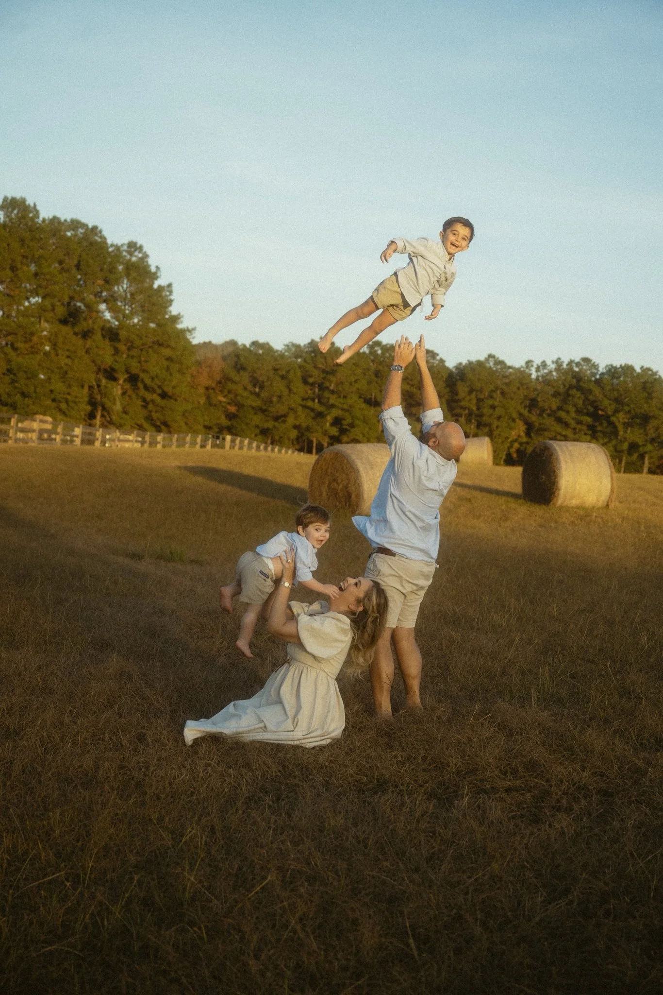 A family in a field holding hands and lifting a young boy into the air, with hay bales and trees in the background during sunset.