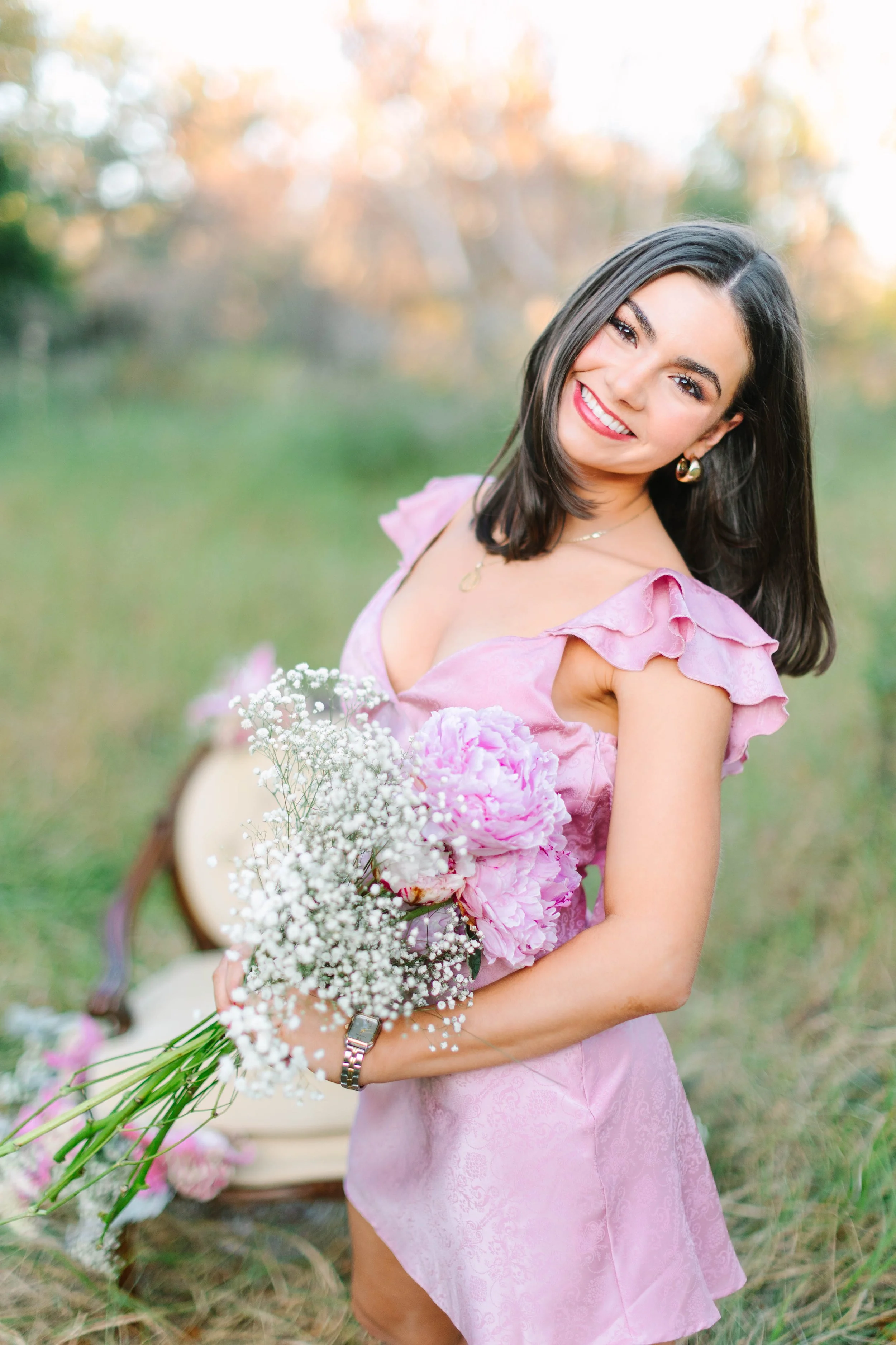 A woman with long dark hair smiling outdoors while holding a bouquet of pink and white flowers. She is wearing a pink dress with ruffled sleeves, gold earrings, and a watch, with a blurred background of trees and grass.
