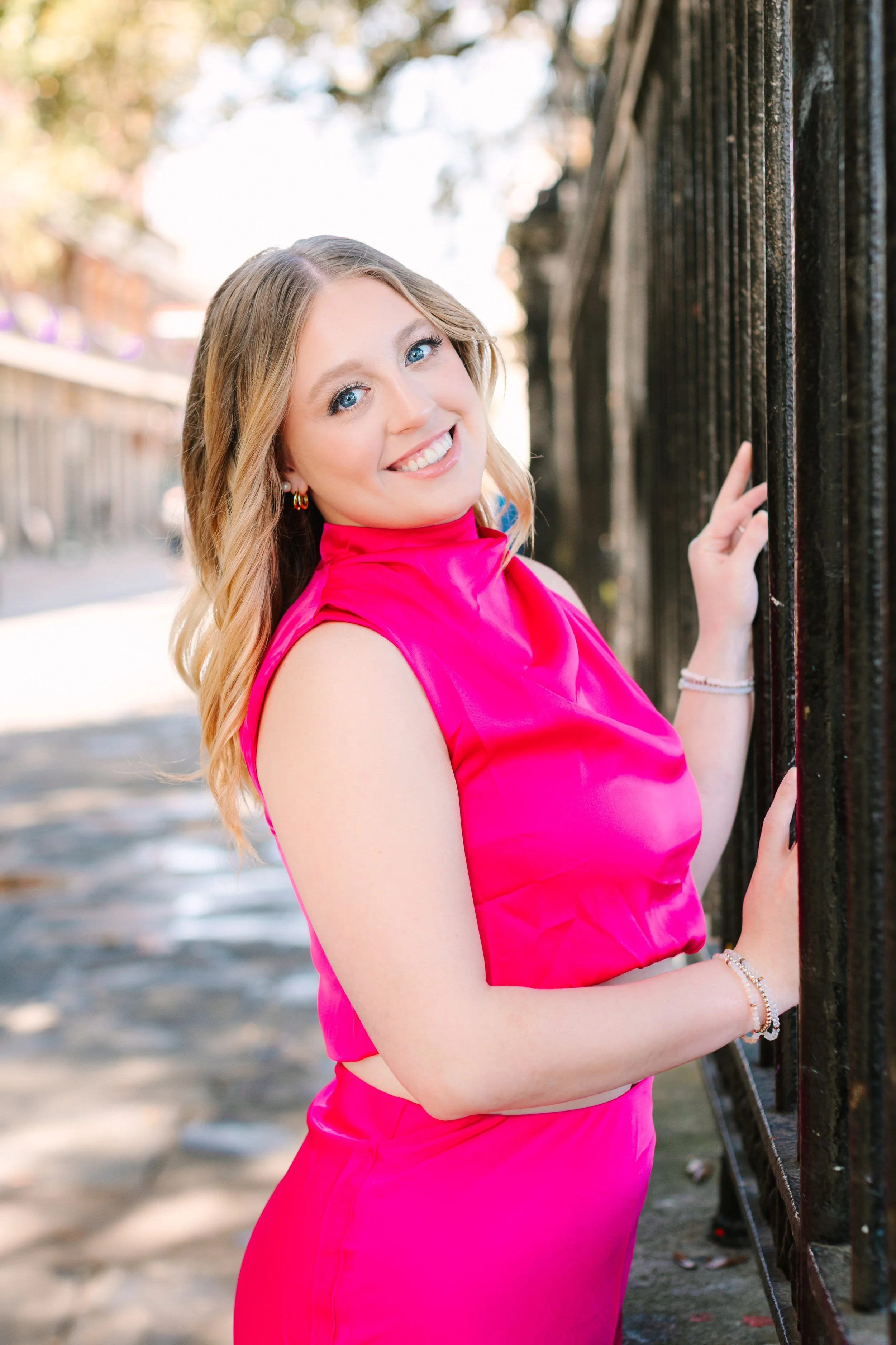 A young woman with blonde hair, wearing a bright pink sleeveless top, standing outdoors and smiling while holding onto a black metal fence.
