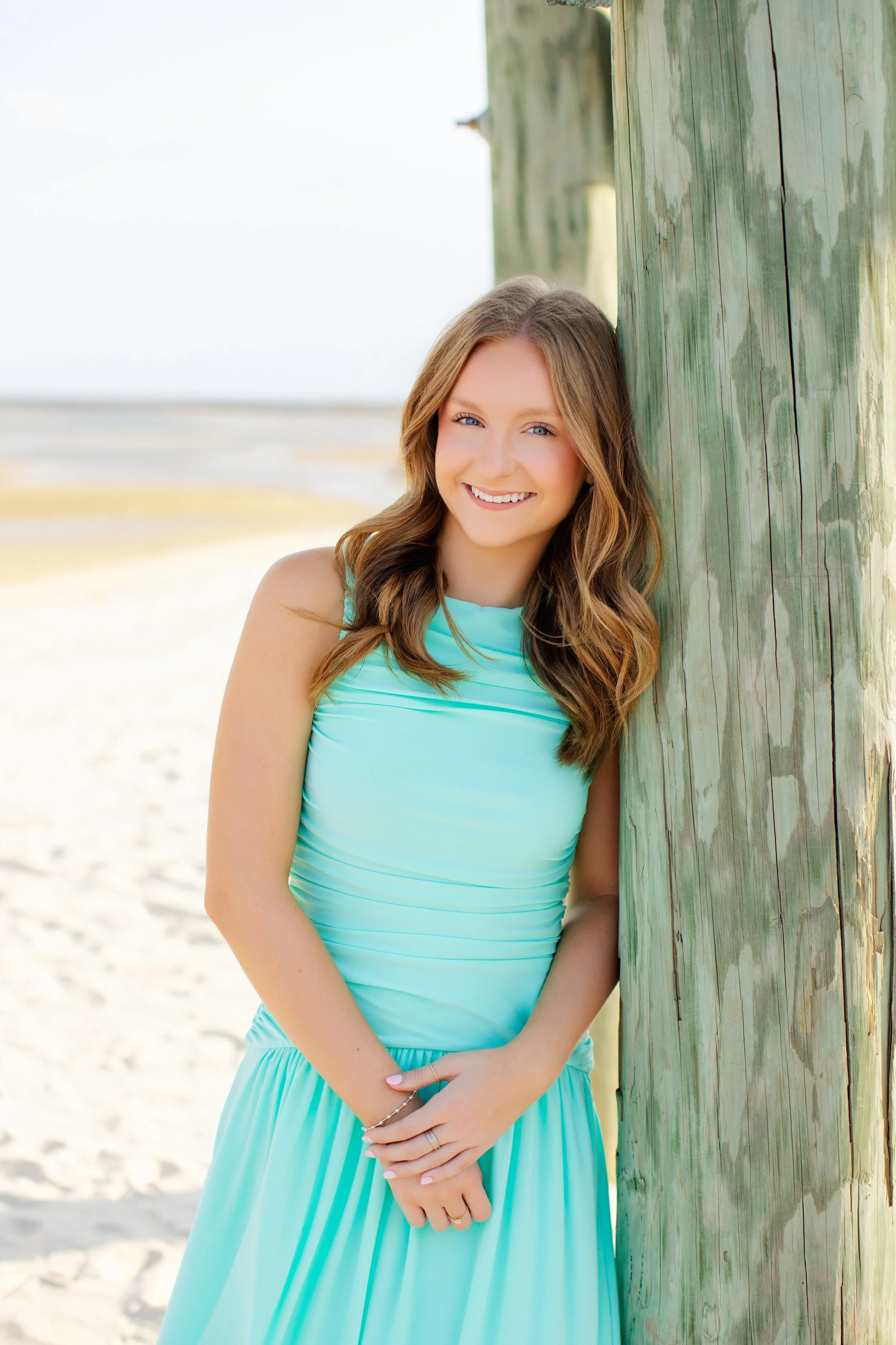 A young woman with long, wavy brown hair smiling and leaning against a large wooden pole on the beach, wearing a light blue sleeveless dress.
