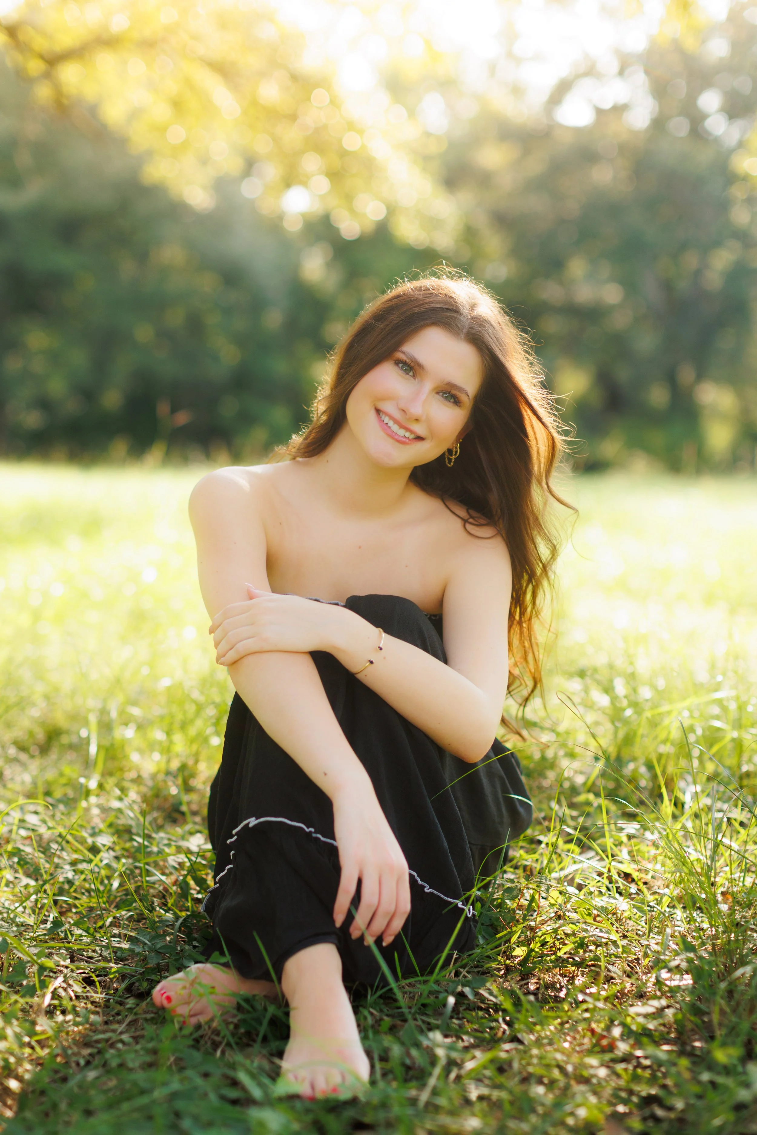 A woman with long brown hair and a bright smile sitting in a grassy outdoor setting, surrounded by trees with sunlight filtering through the leaves.
