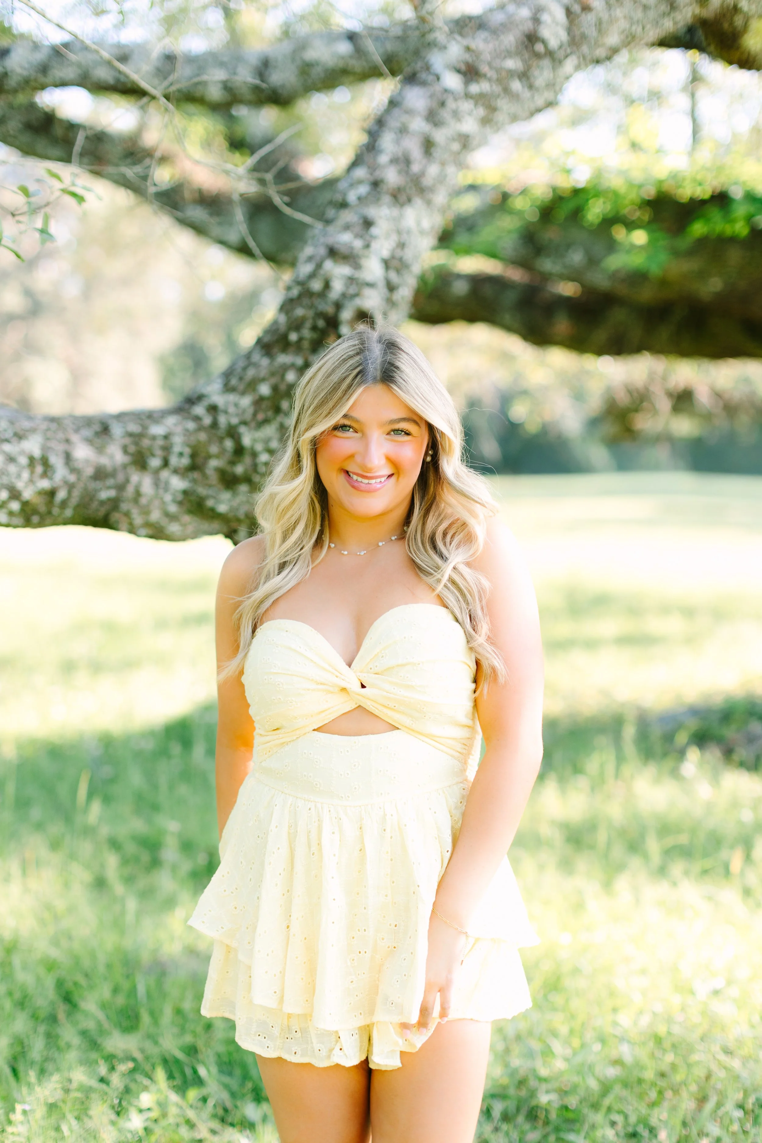 A young woman with long blonde hair wearing a light yellow strapless dress standing outdoors in a grassy area with a large tree in the background.