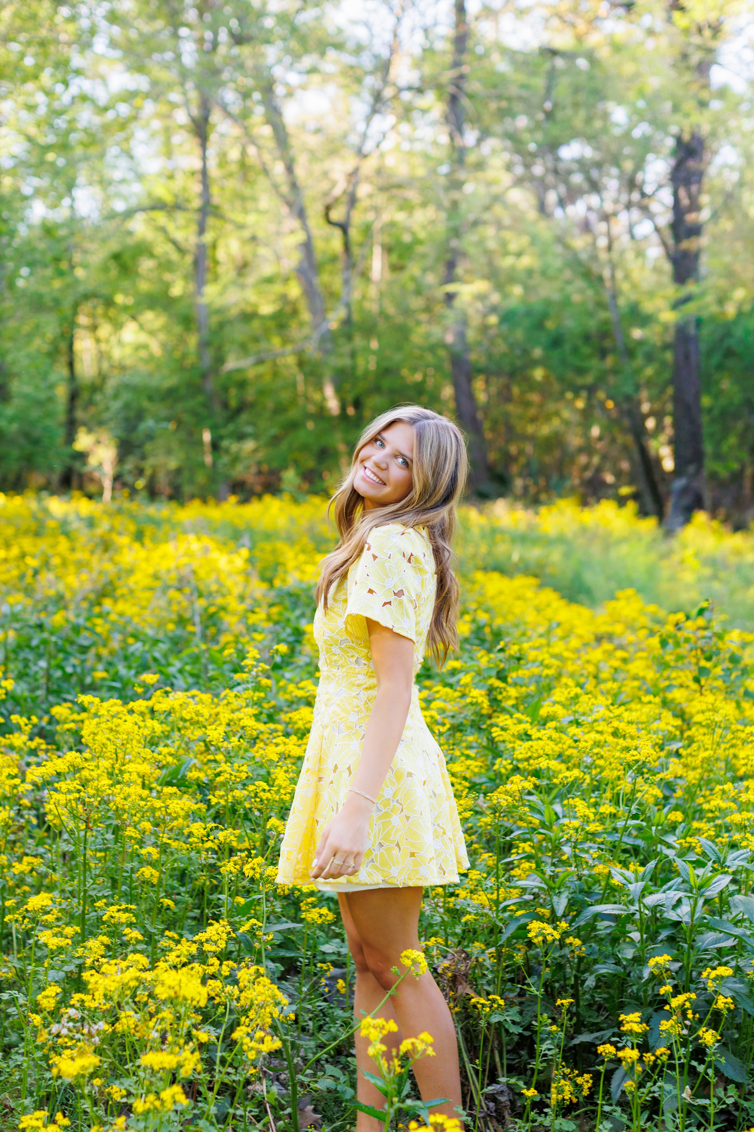 A smiling woman in a yellow dress standing in a field of yellow flowers with a wooded area in the background.